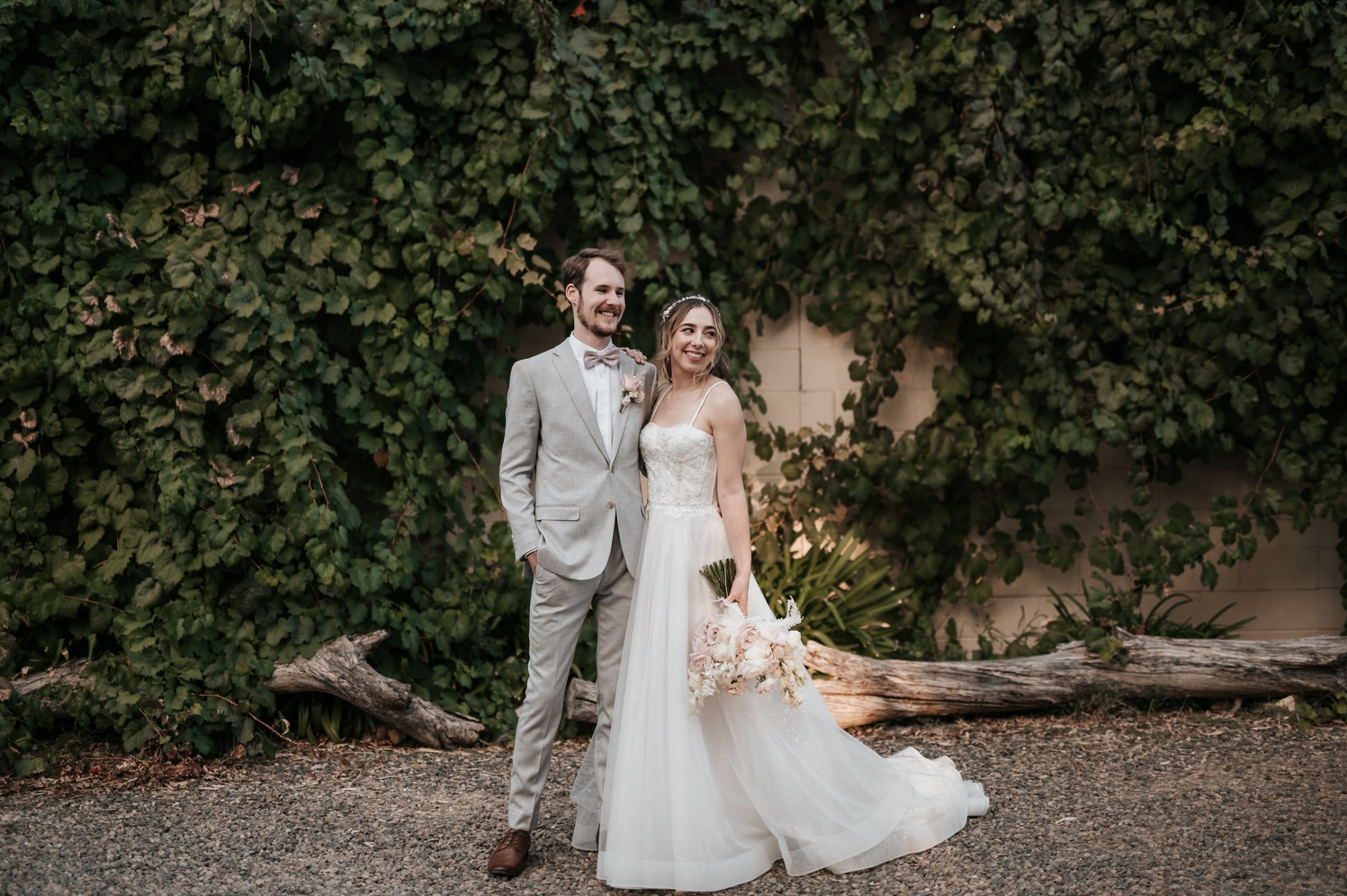 Smiling wedding couple posing outdoors in front of a lush green vine-covered wall.