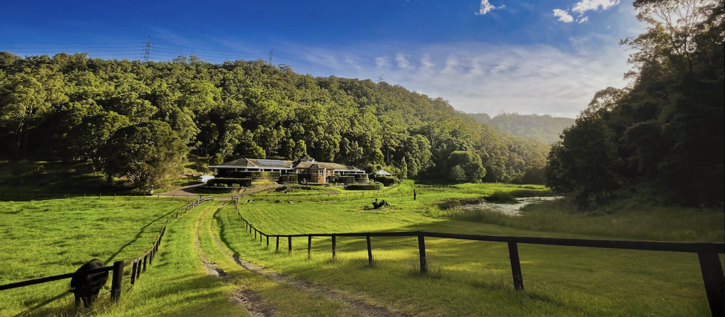 Scenic countryside wedding venue with a farmhouse, rolling green fields, and forested hills under a blue sky.