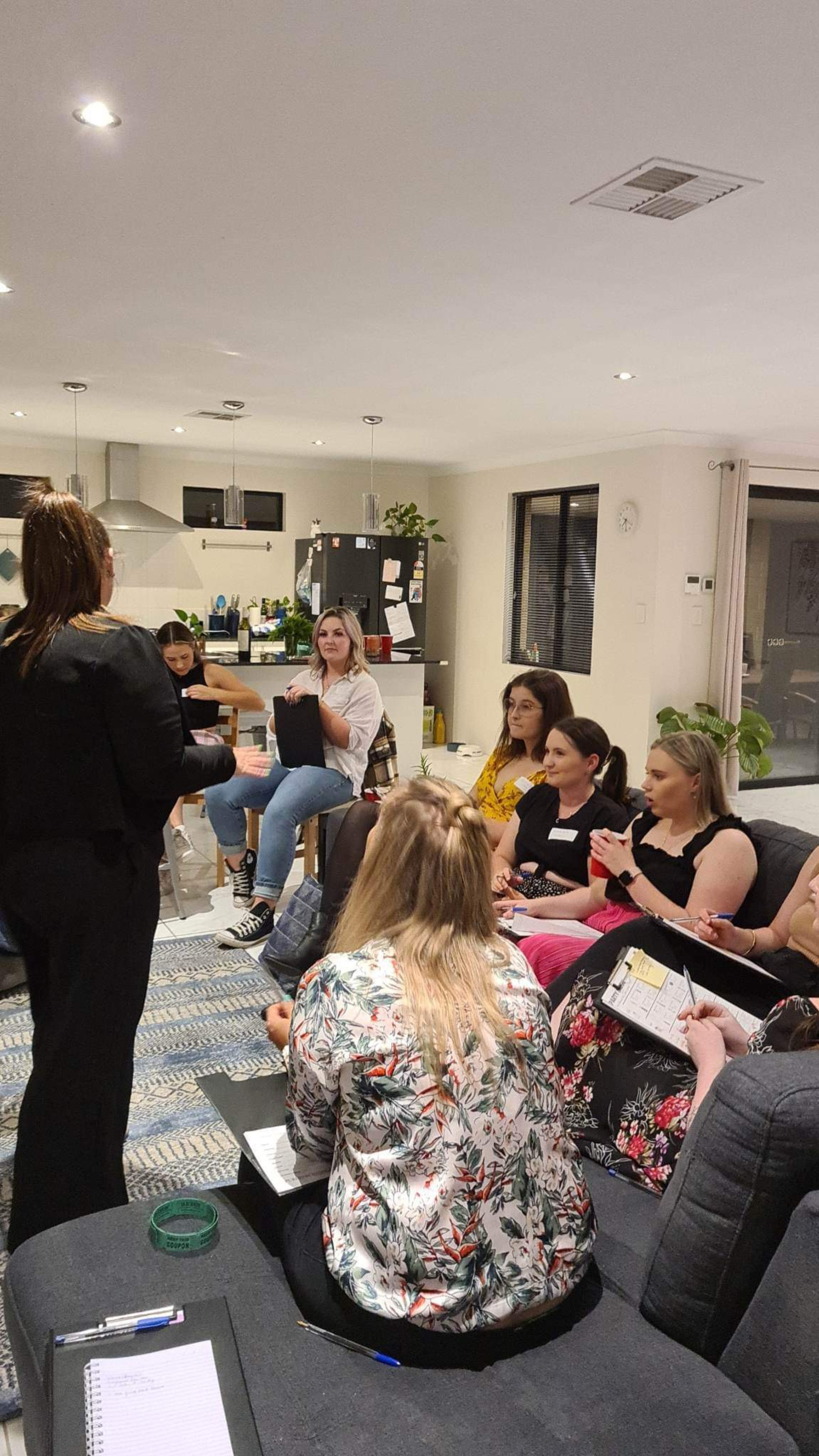 A group of women sit in a modern living room taking notes during a wedding planning workshop.