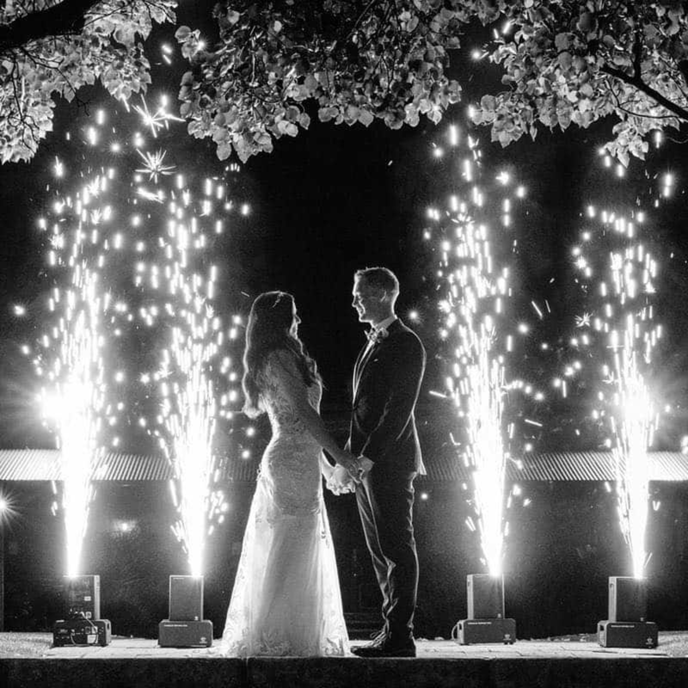 Bride and groom hold hands at night surrounded by tall indoor fireworks and sparkling lights.