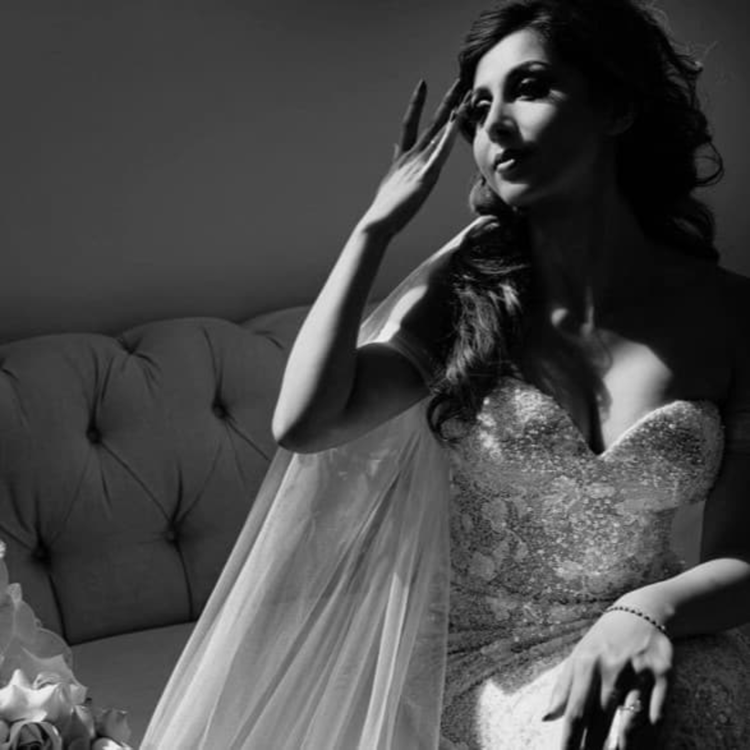 Black and white portrait of a bride in a sparkly gown sitting on a sofa, softly lit as she touches her hair.