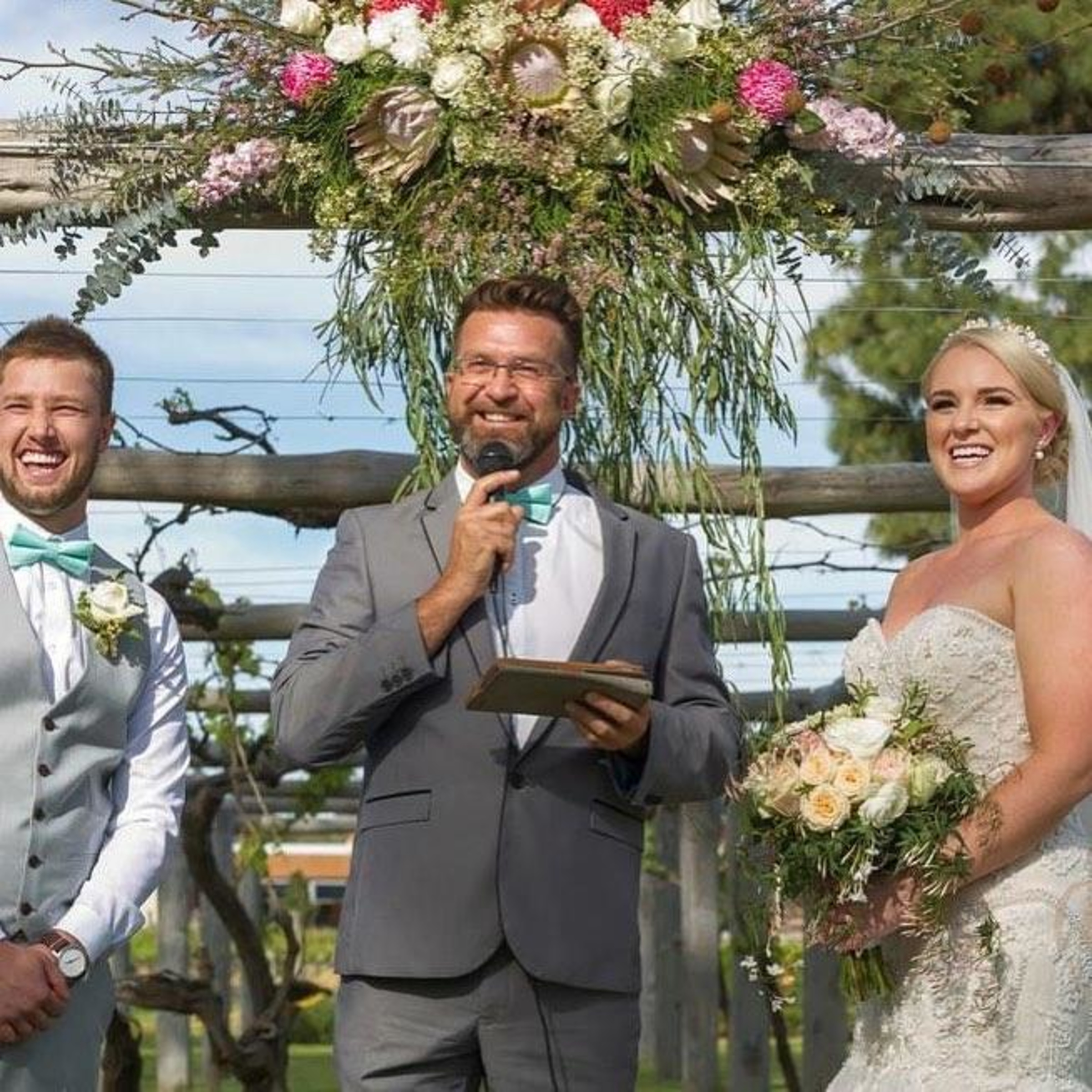 Outdoor vineyard wedding ceremony with celebrant speaking beside smiling bride and groom under floral arch.