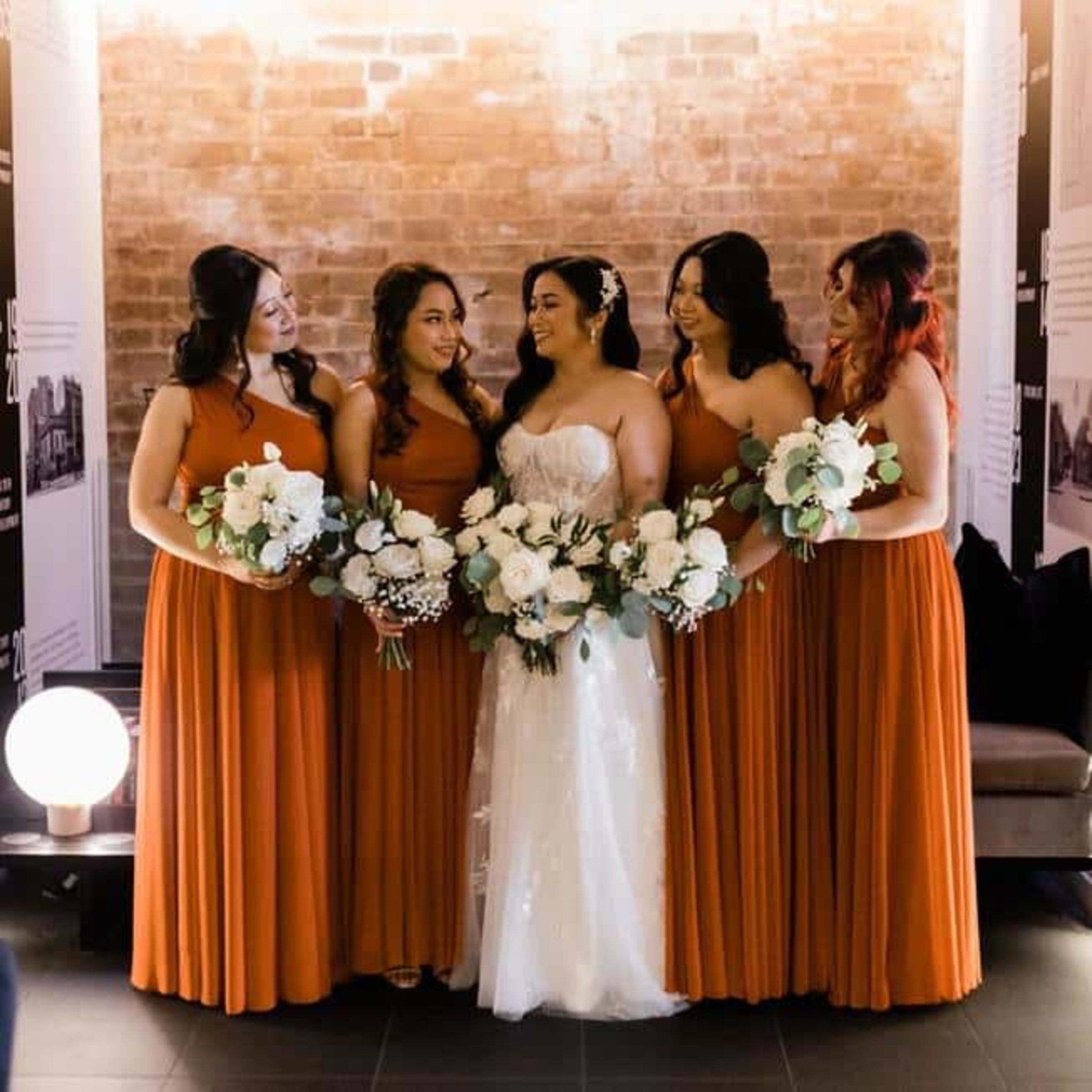 Bride in a white gown stands with bridesmaids in rust dresses holding white and green bouquets against a brick wall.