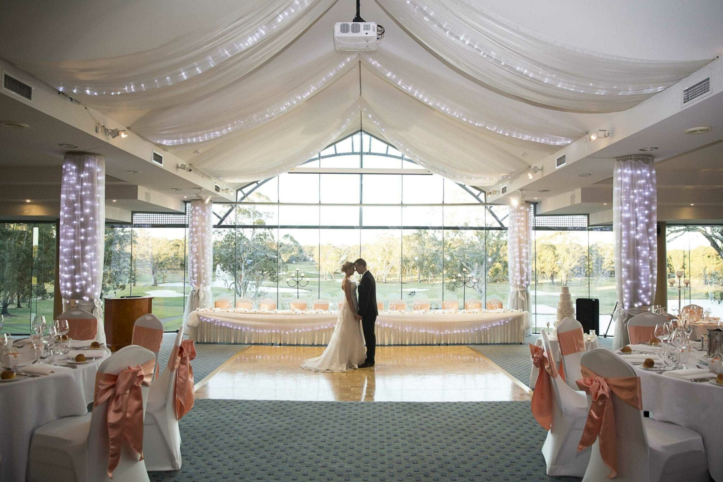 Bride and groom share a moment on the dance floor in a light-filled wedding reception hall with draped ceilings and peach accents.