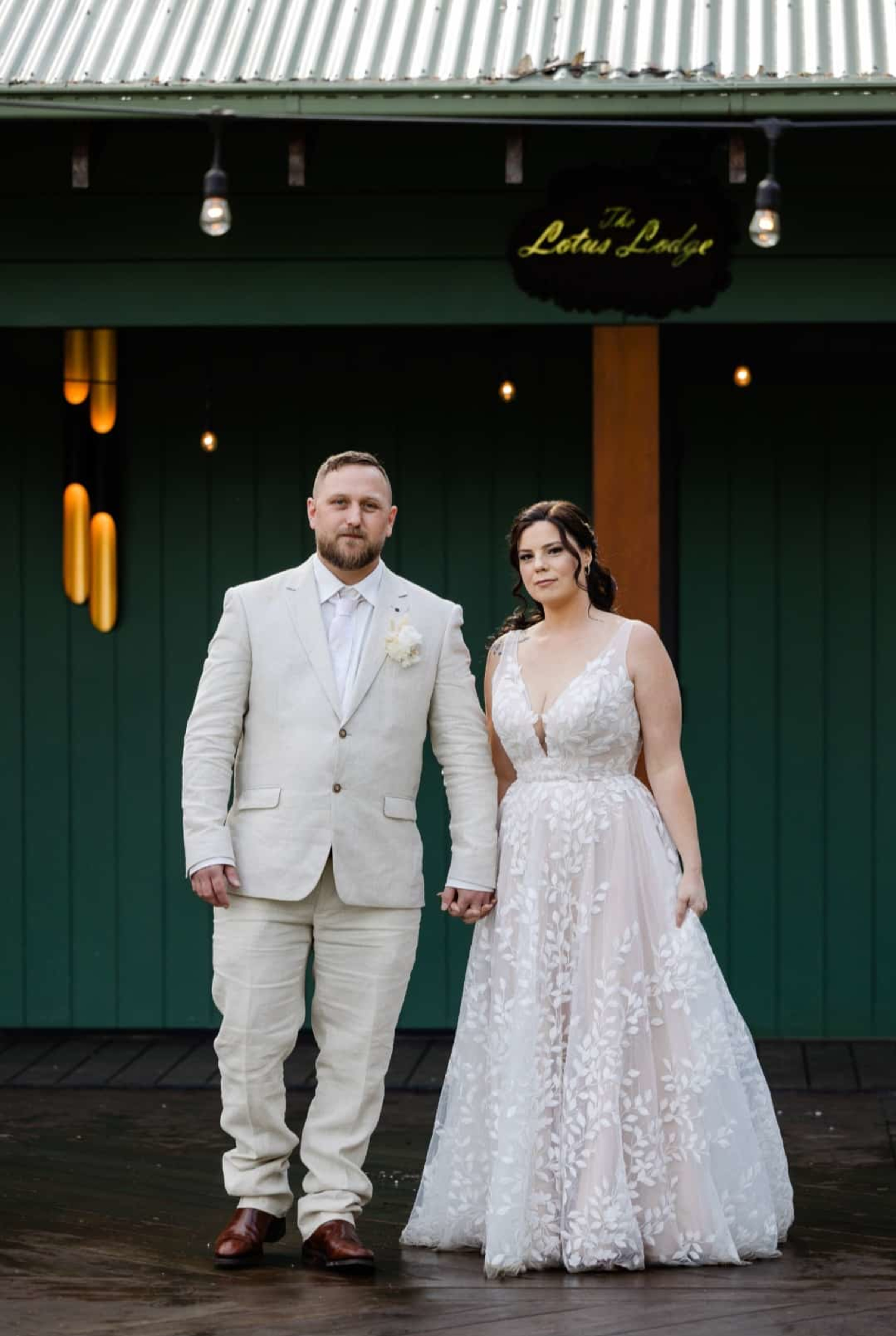 Bride and groom stand holding hands in front of a green lodge backdrop with warm hanging lights.