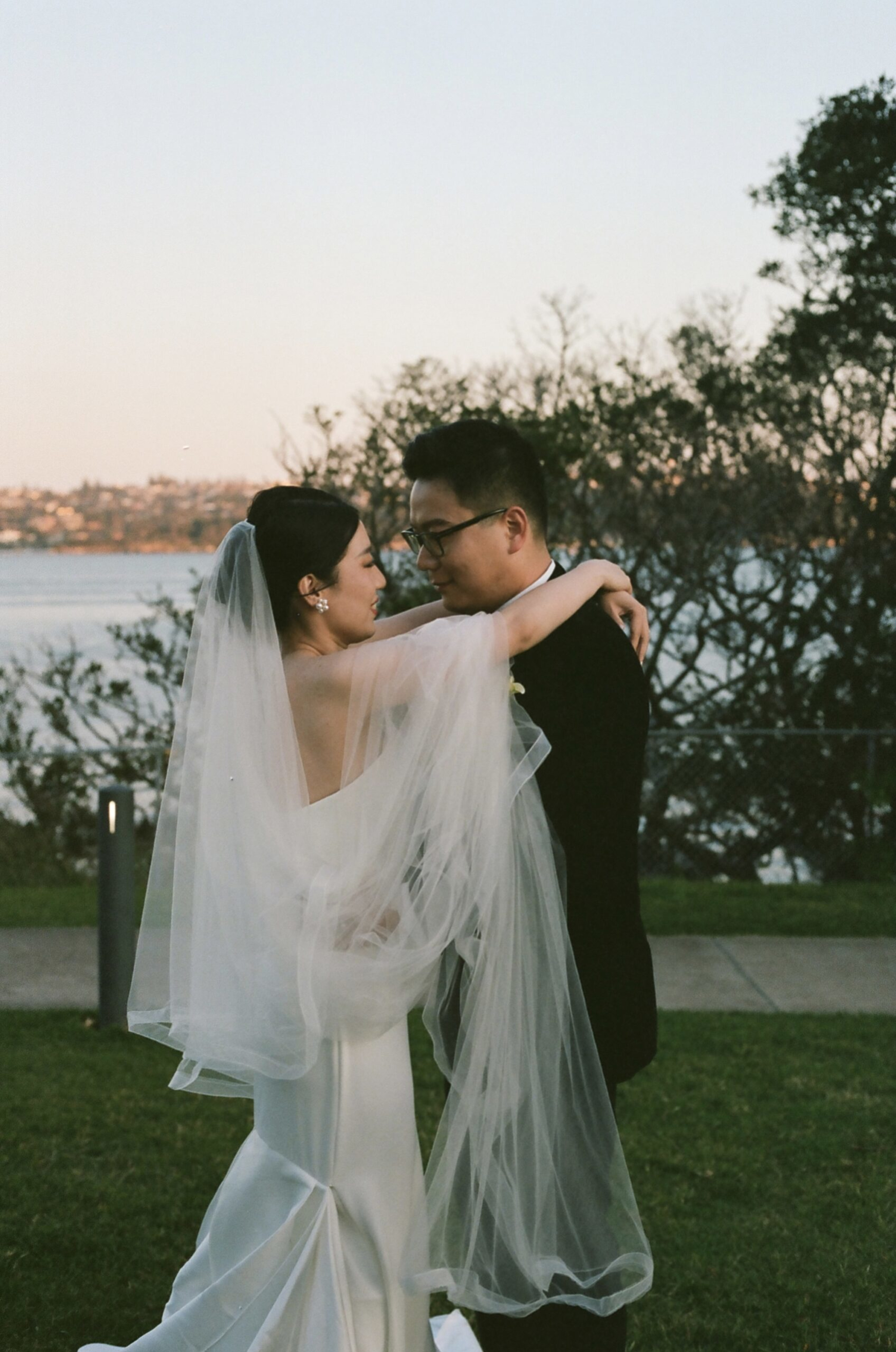 Bride and groom embrace in an outdoor waterfront setting at sunset, with her veil flowing around them.