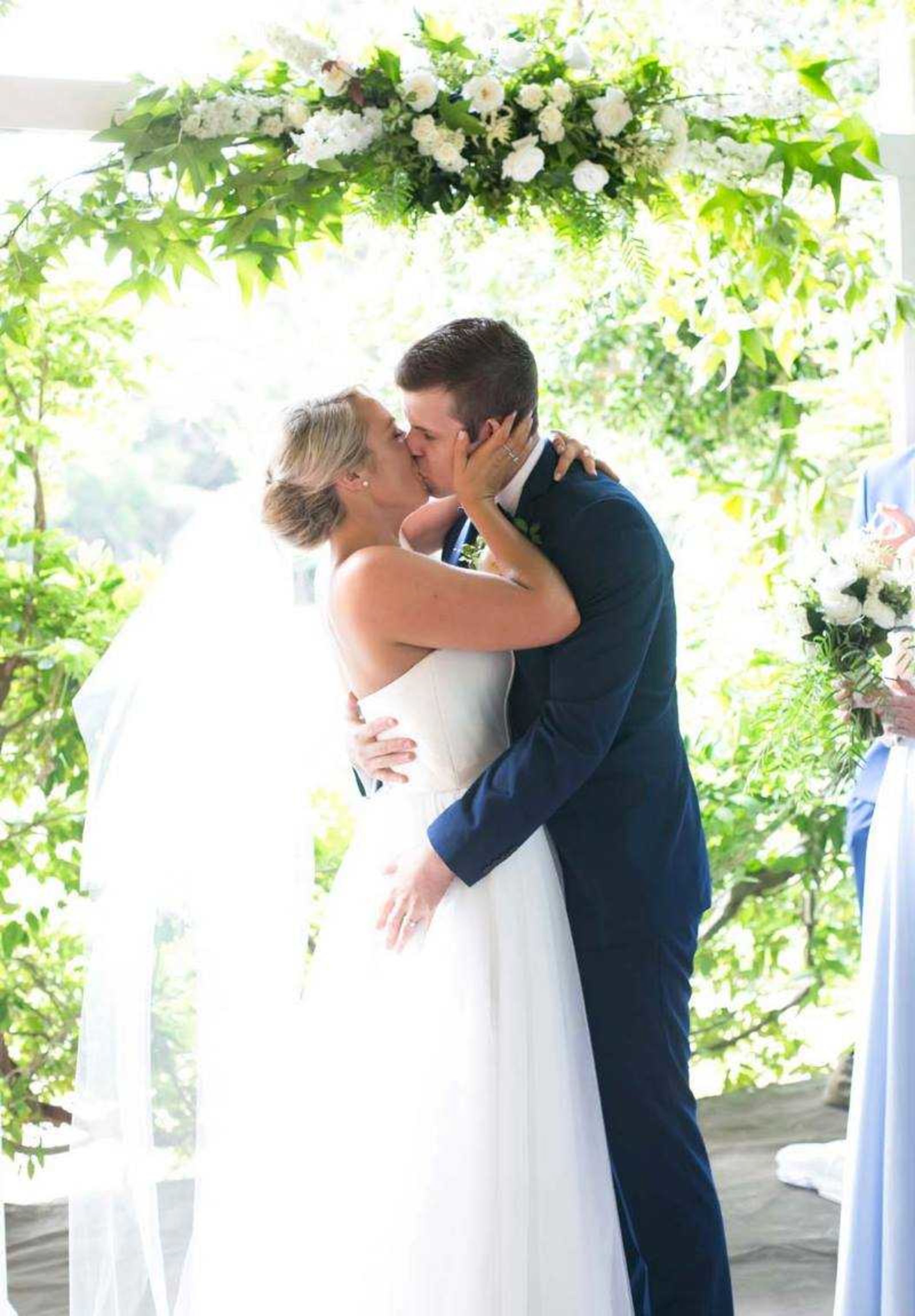 Bride and groom share a kiss beneath a lush floral arch during their outdoor wedding ceremony.