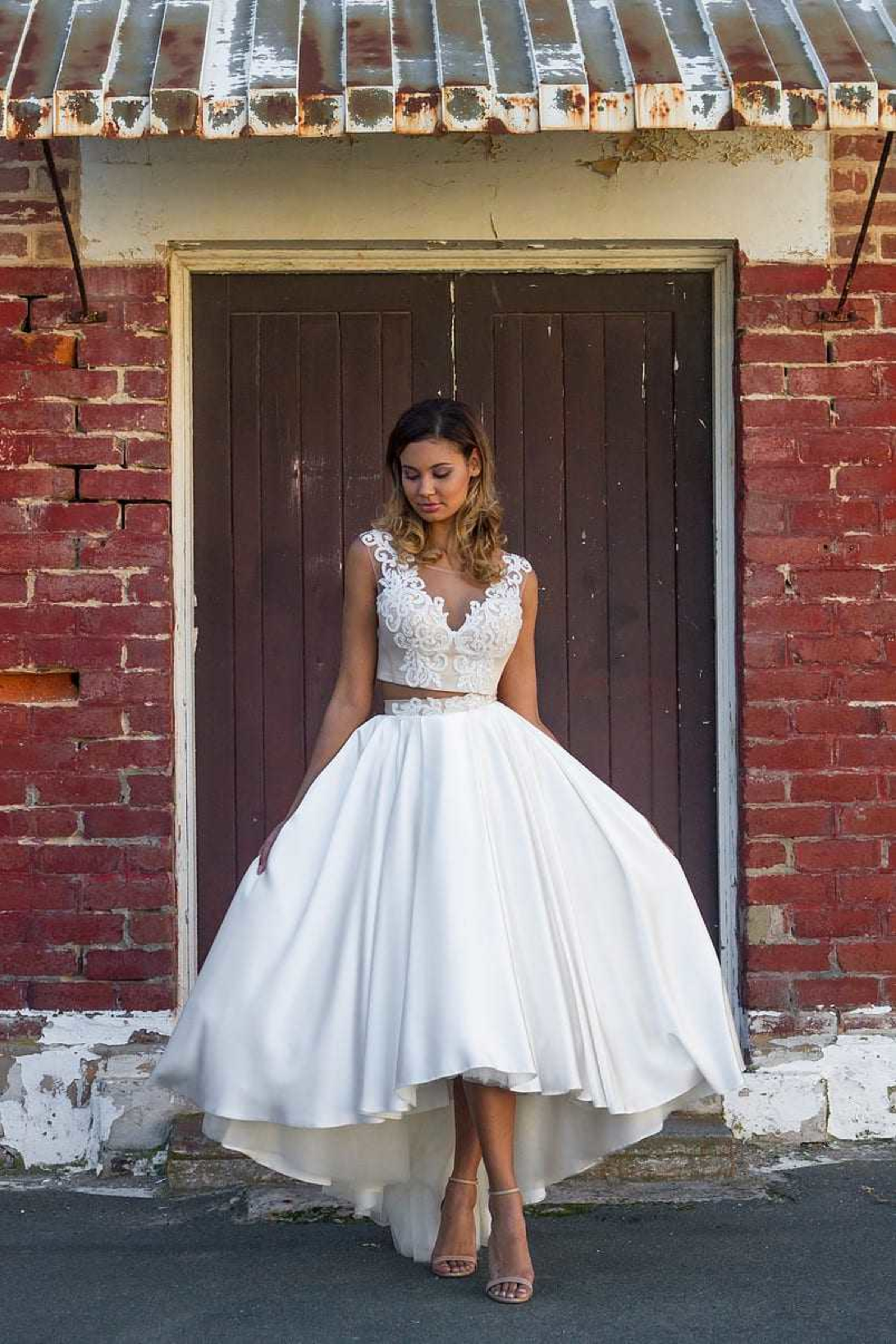 Bride in a high-low lace and satin wedding dress posing against a rustic brick wall and wooden door.