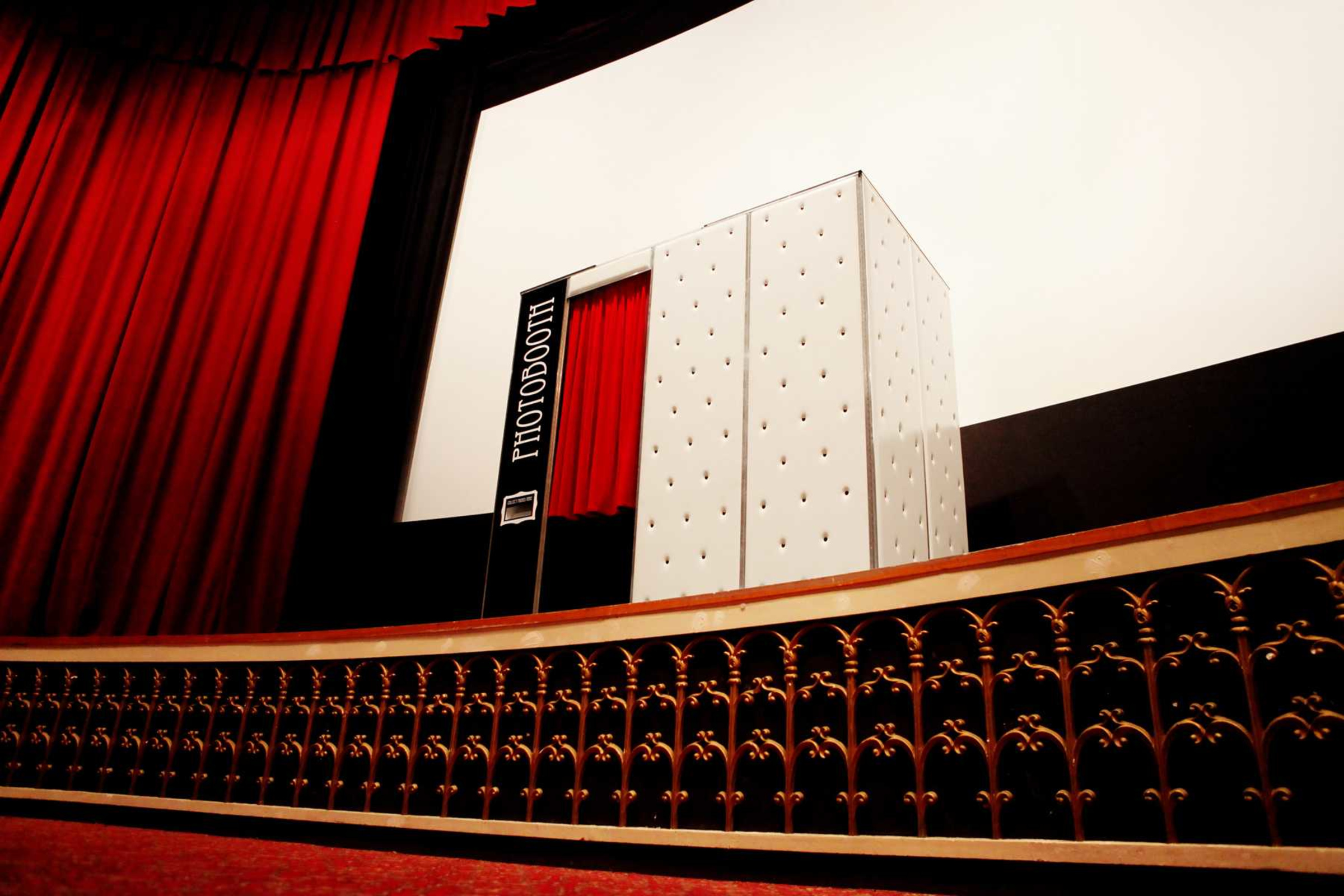 Elegant white photobooth with red curtain set on a theater-style stage with dramatic red drapes.