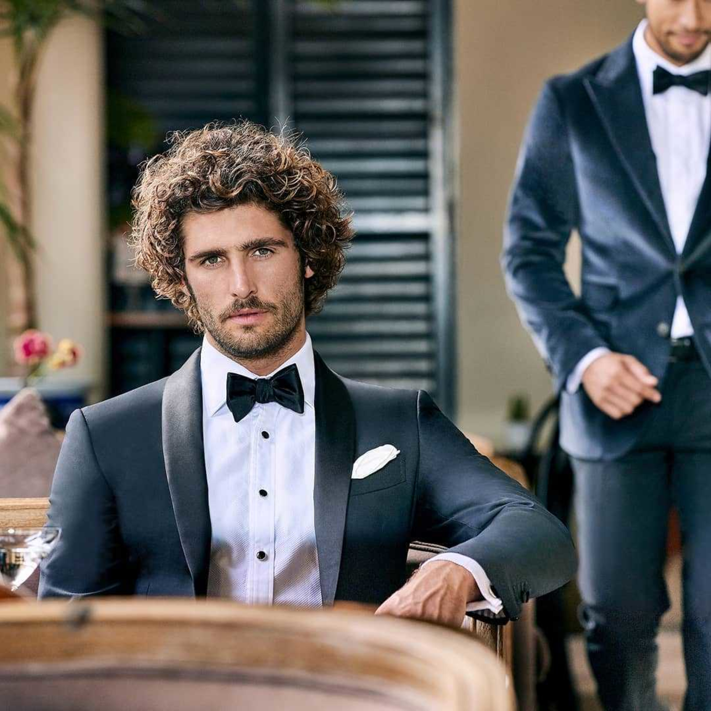Stylish groom and groomsman in black tuxedos posing in an elegant indoor wedding setting.