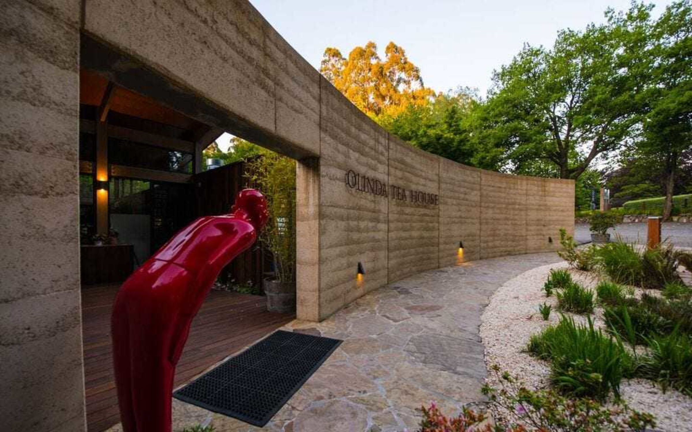 Modern tea house entrance with curved stone wall, red sculpture, and landscaped garden path at sunset.