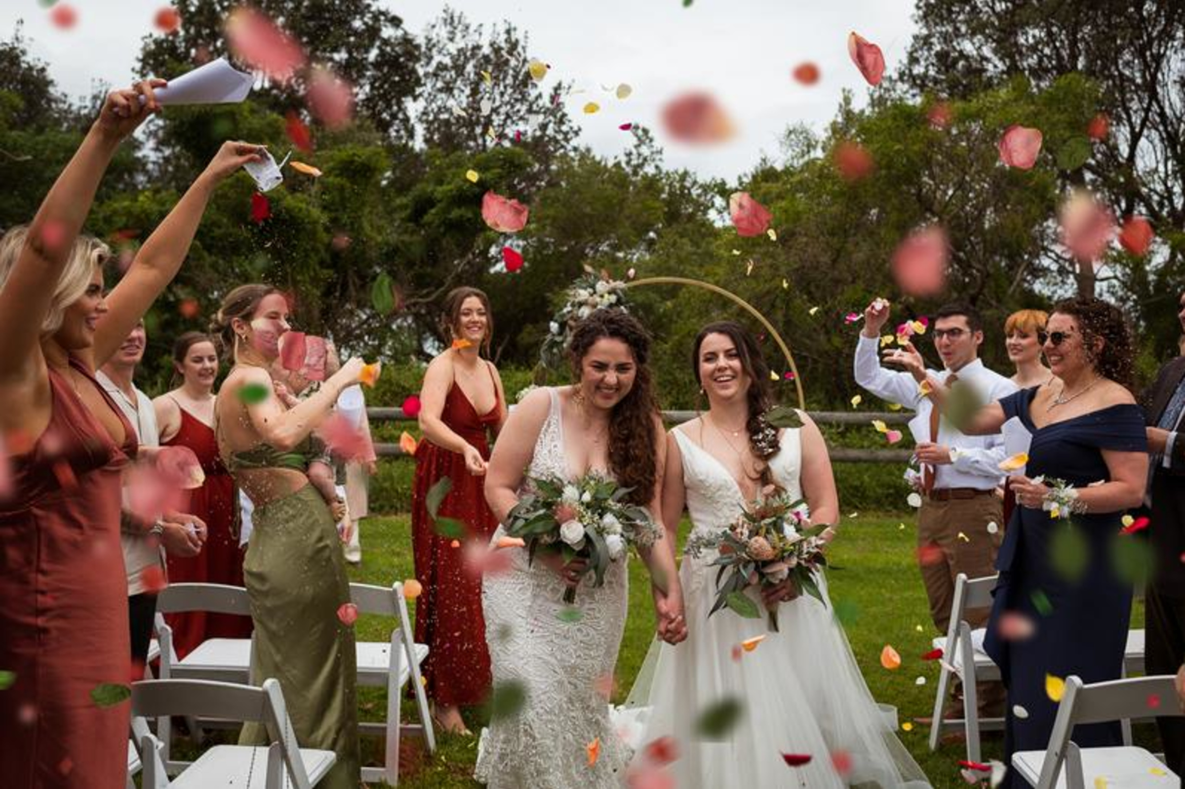 Two brides walk hand in hand down the aisle as guests toss colorful petals during an outdoor wedding celebration.