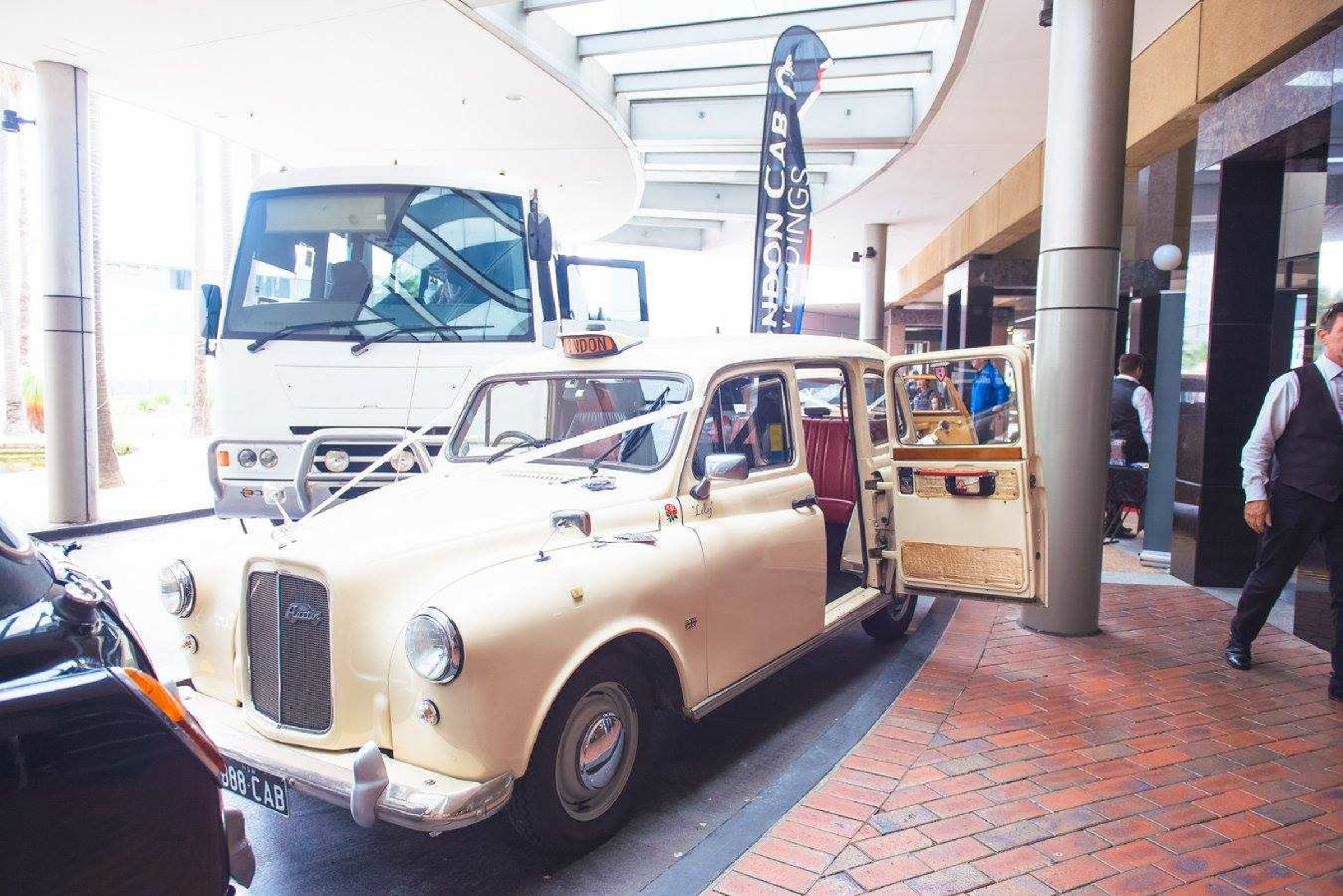 Cream vintage London taxi decorated with wedding ribbons parked at a modern city venue entrance.