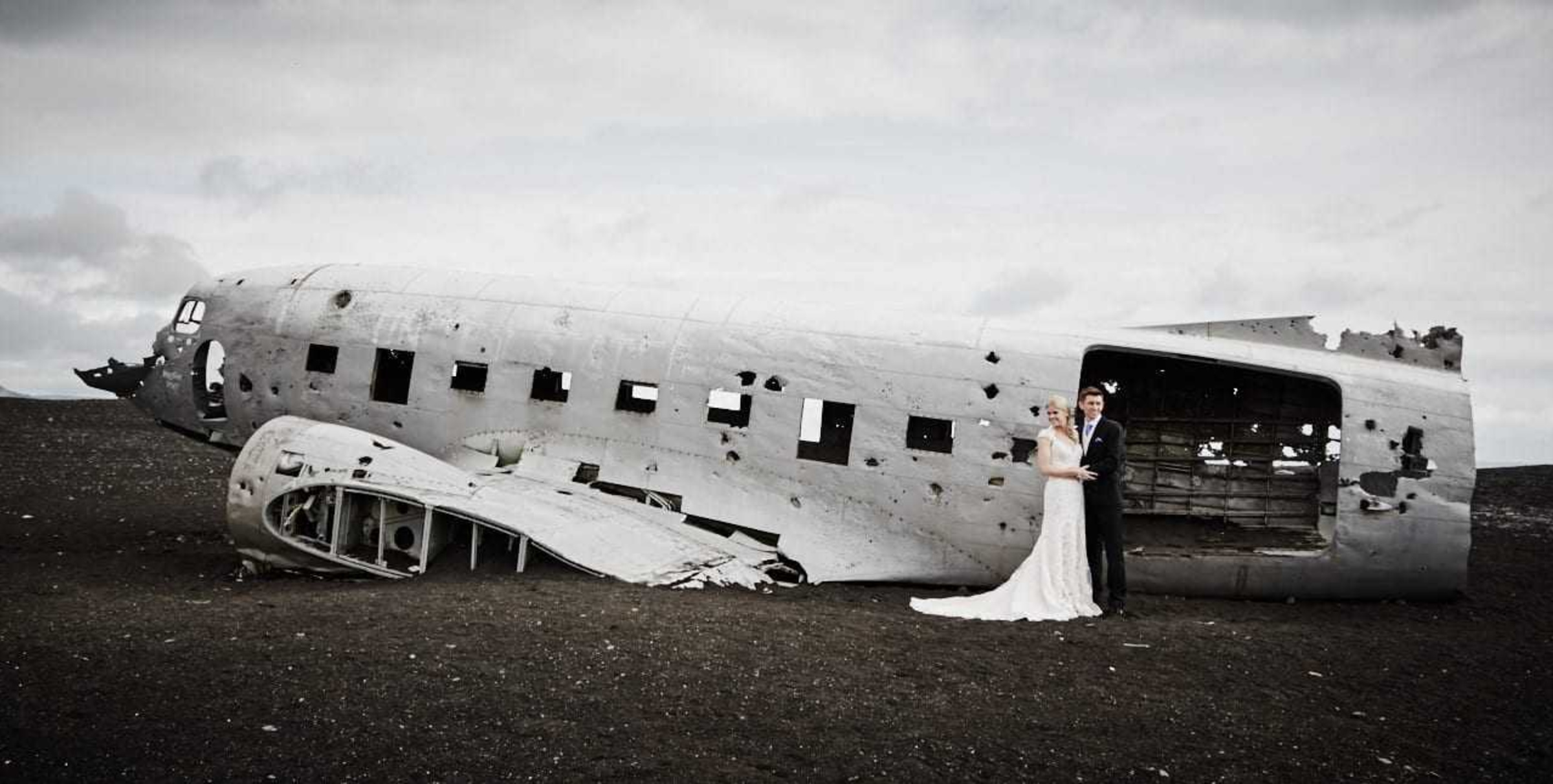 Bride and groom posing beside a dramatic crashed airplane wreck in a barren landscape under cloudy skies.