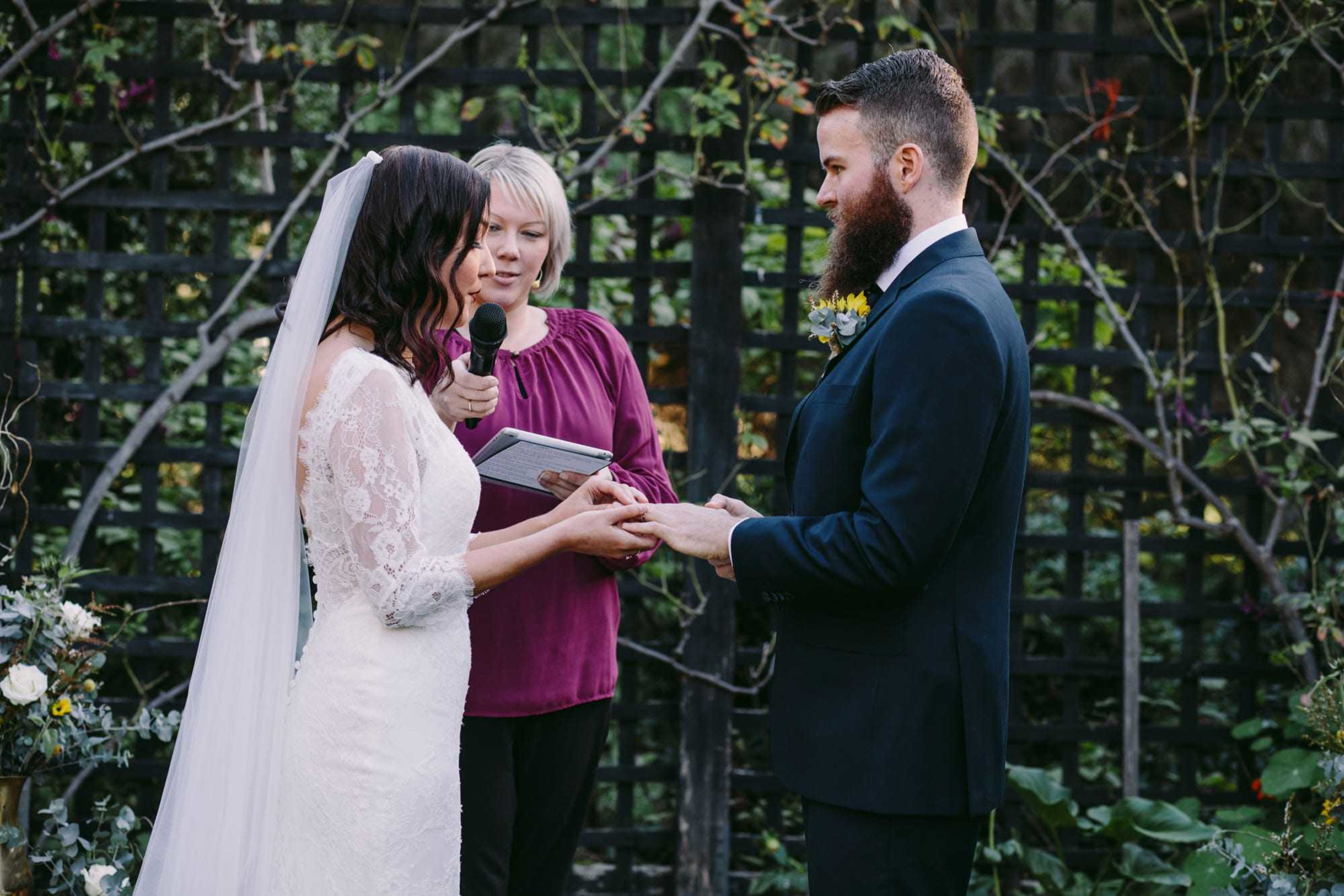 A couple exchanges vows during an intimate outdoor wedding ceremony, officiated by a woman holding a microphone and tablet.