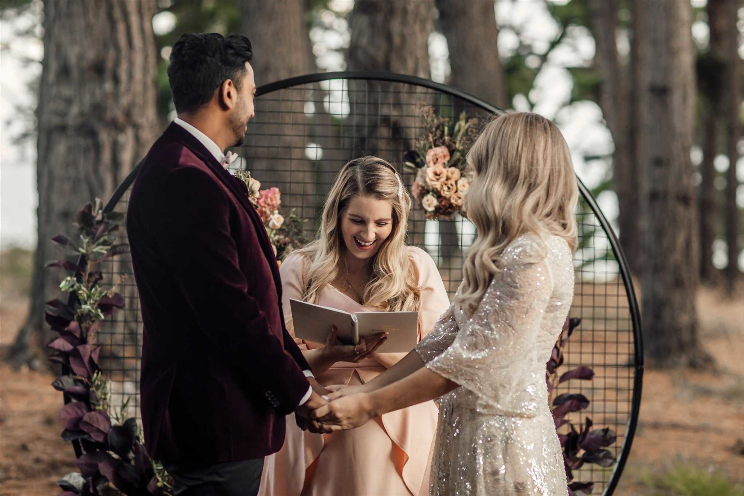 An officiant leads an outdoor forest wedding ceremony as the couple holds hands beneath a circular floral arbor.