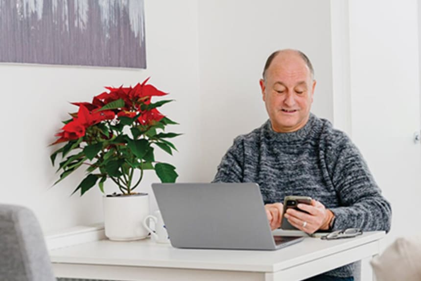 Older man sitting in front of a laptop while also looking at his phone