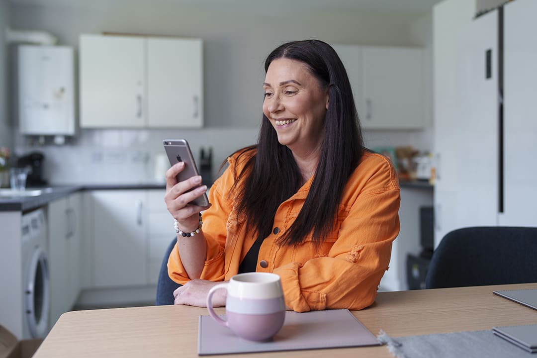 Female customer smiles while she uses My Sage Home online account on her phone, sat in her kitchen