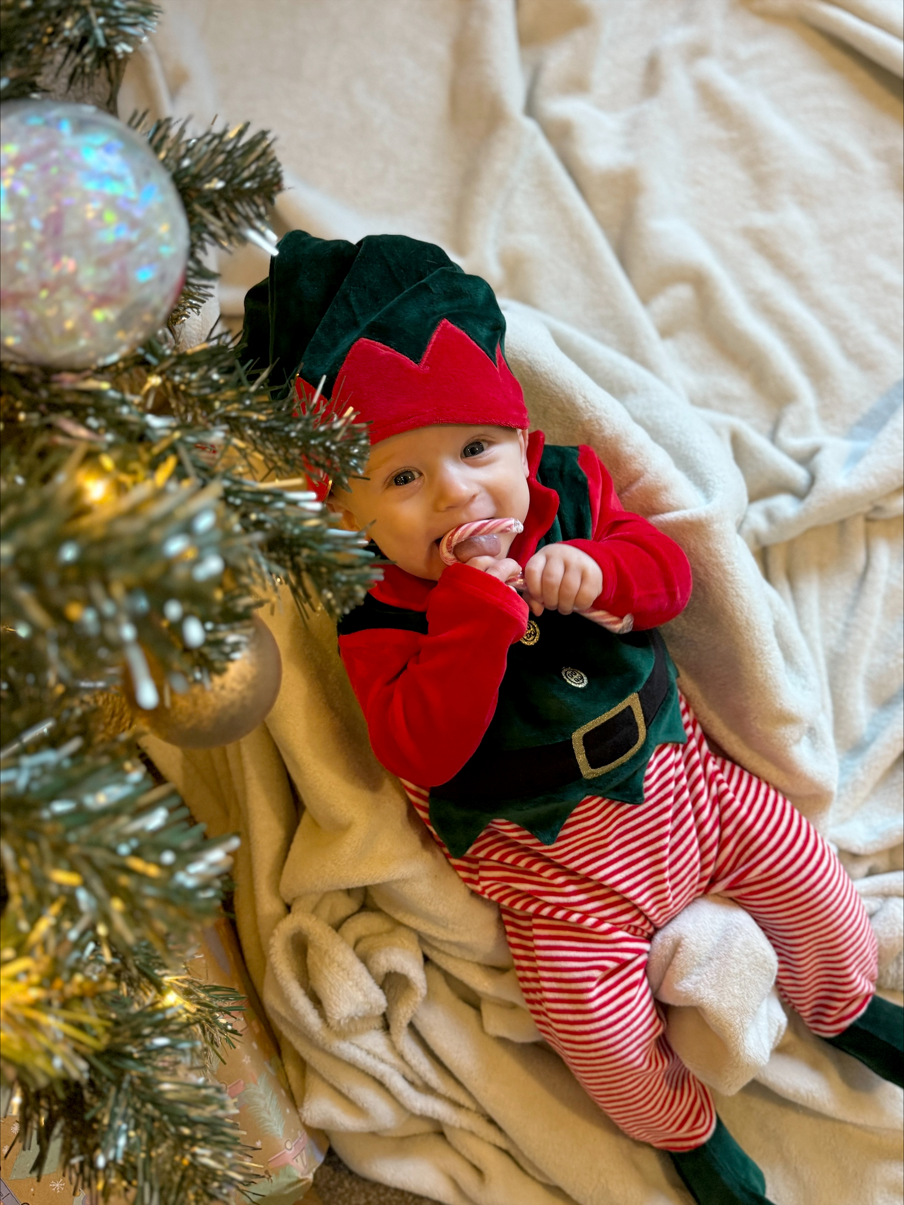 A cute baby in an elf outfit, under a Christmas tree