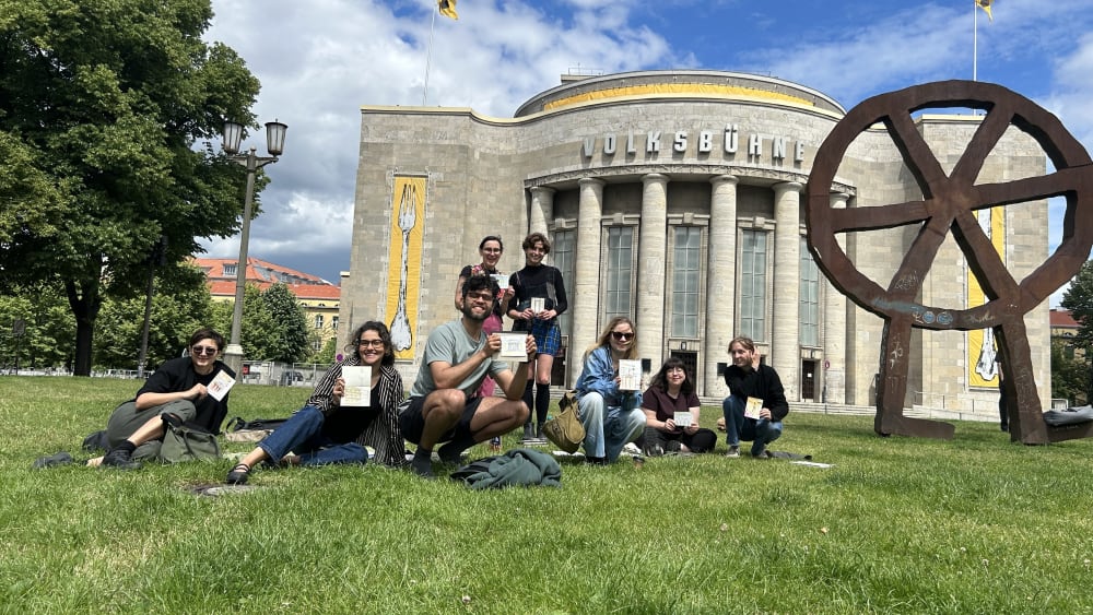 A group sitting on the grass in front of the Volksbühne, each holding up their painting