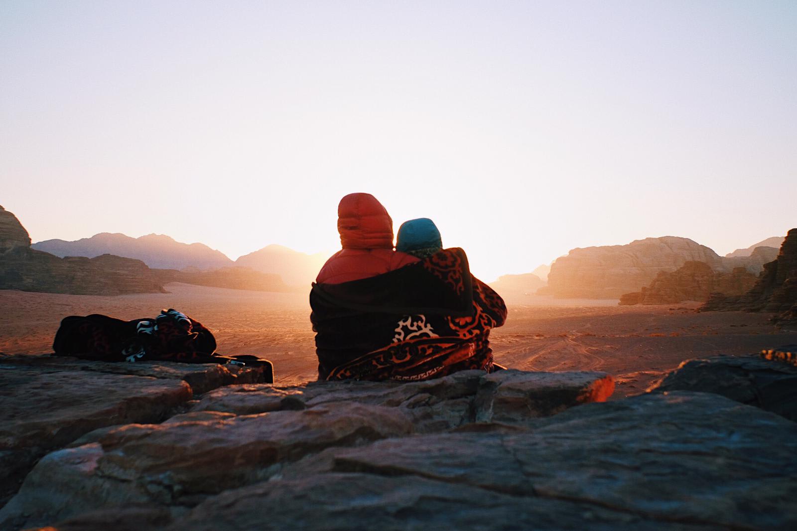Two people in warm clothes sitting on rocky ground, watching the sunrise in a desert landscape.