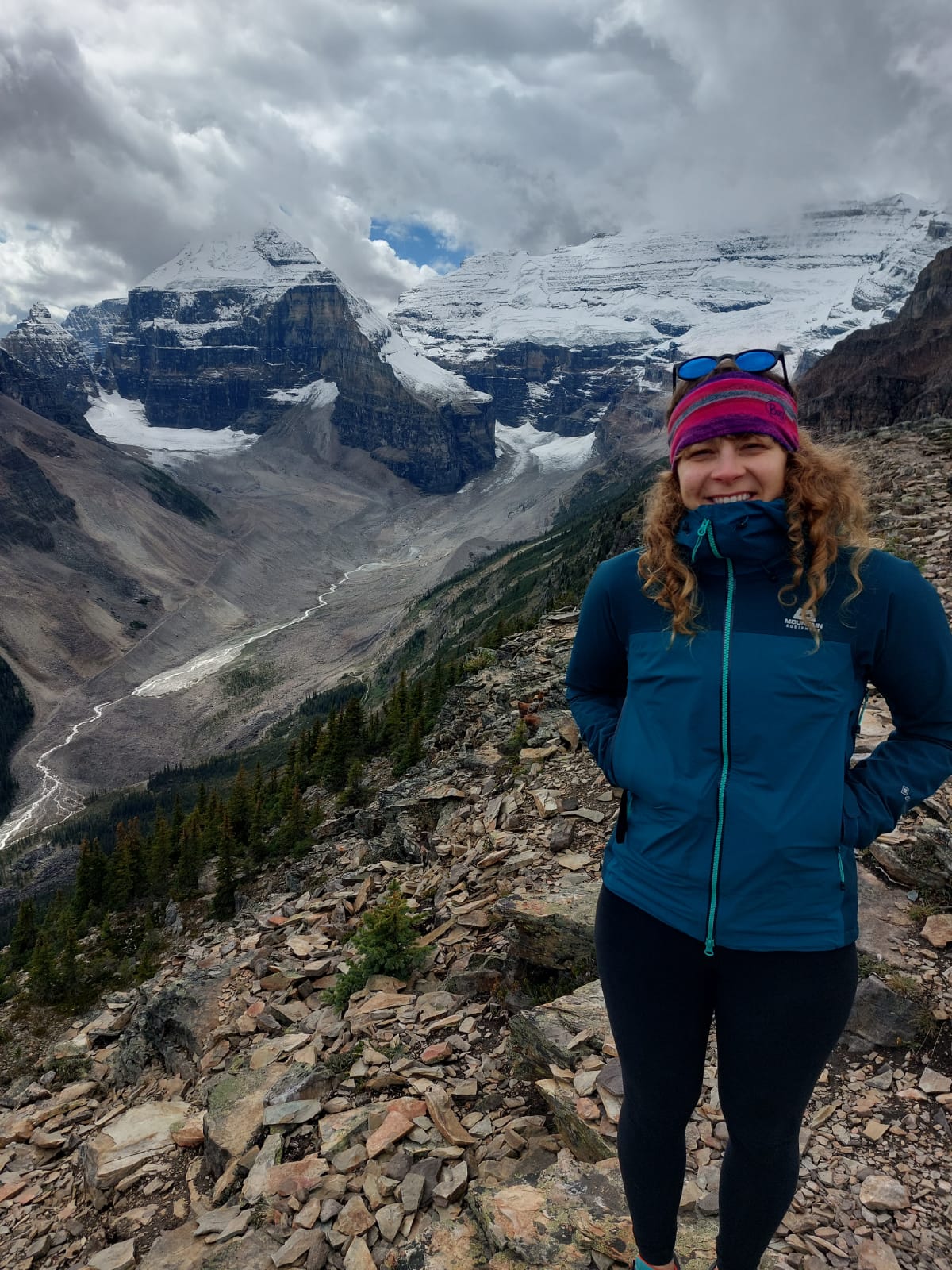 A woman in a blue jacket and pink hat standing on a rocky mountain trail with snow-capped peaks in the background.