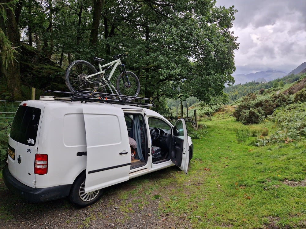 White van with a bicycle on its roof rack parked on a grassy area near trees, with mountains visible in the distance.