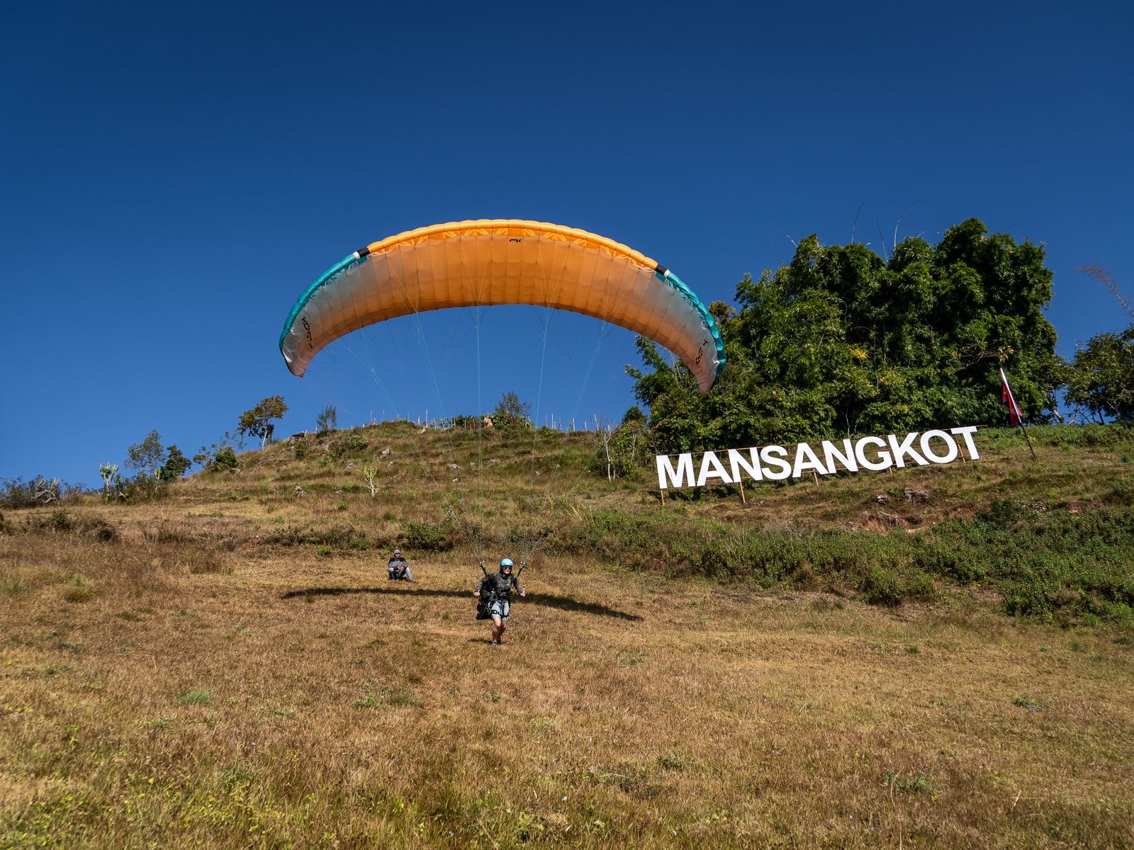 A paraglider with a yellow and blue parachute prepares to land on a grassy hill marked "MANSANGKOT" under a clear blue sky.