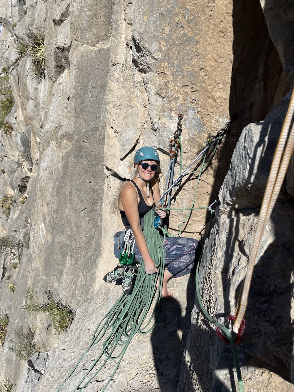 Person rock climbing with ropes, wearing helmet and sunglasses, mid-climb on a steep rocky surface with climbing gear.