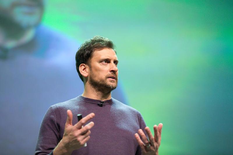 A man with dark hair and beard gestures while speaking on a stage.