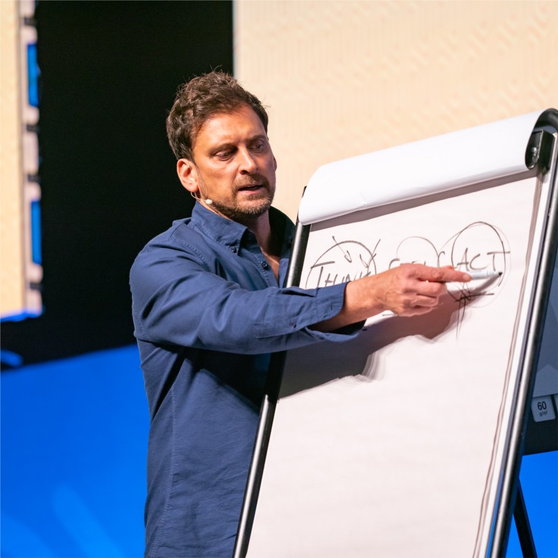 A man in a blue shirt points at a flip chart with diagrams during a presentation.