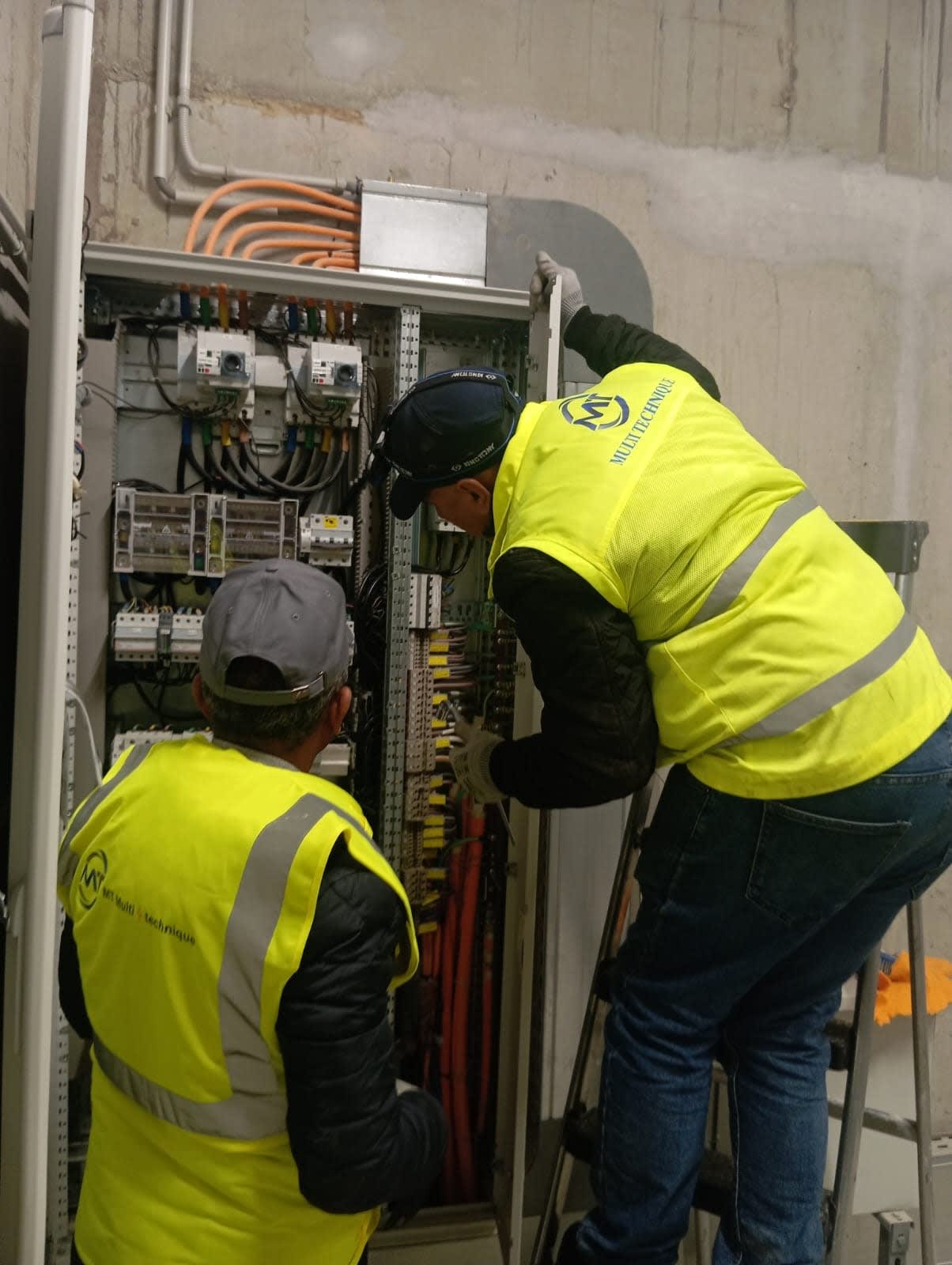 Deux techniciens MT Multi Technique en gilet haute visibilité interviennent dans une armoire électrique TGBT ouverte