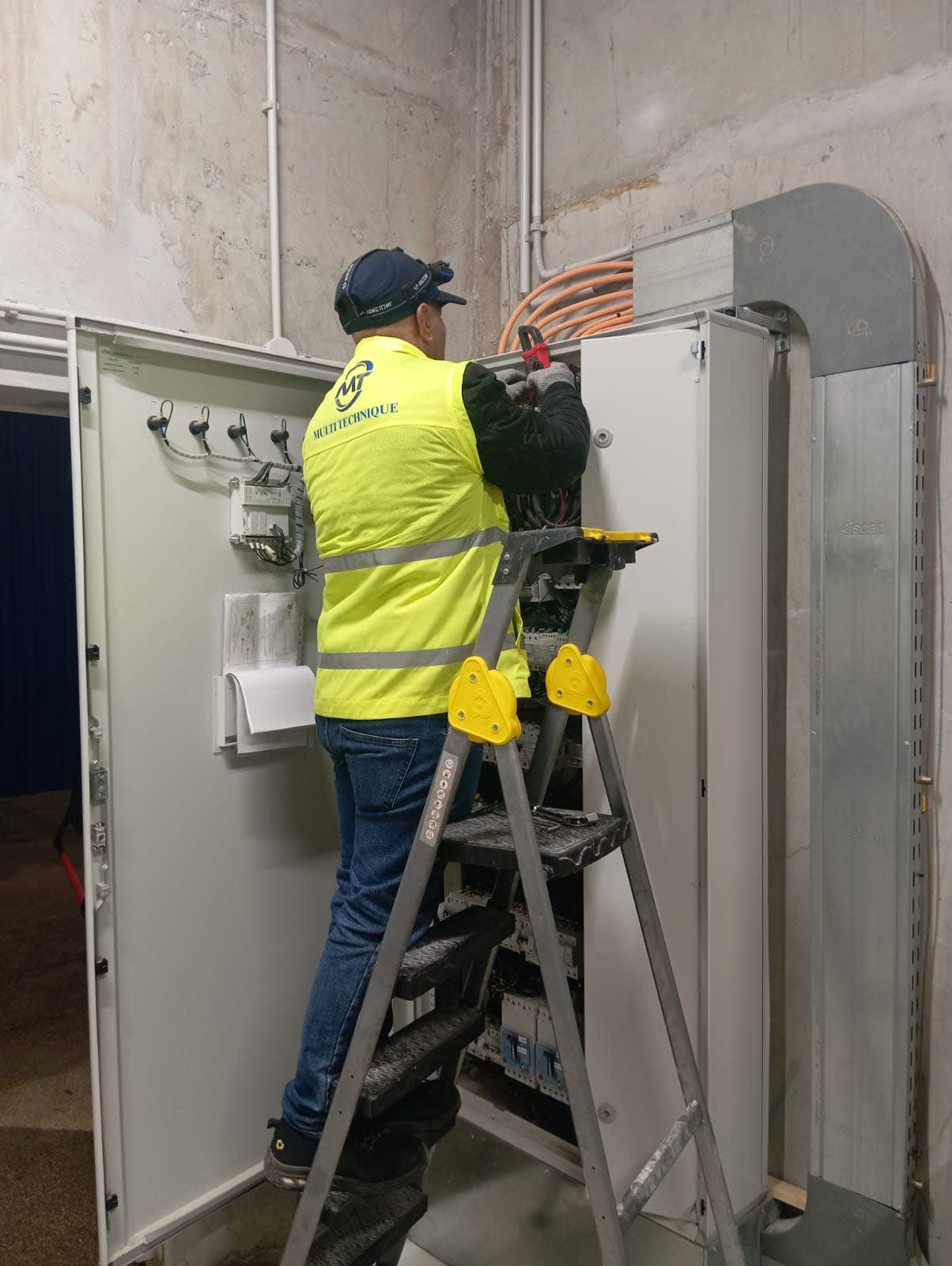 Technicien sur escabeau effectuant une mesure à la pince ampèremétrique dans une armoire de distribution
