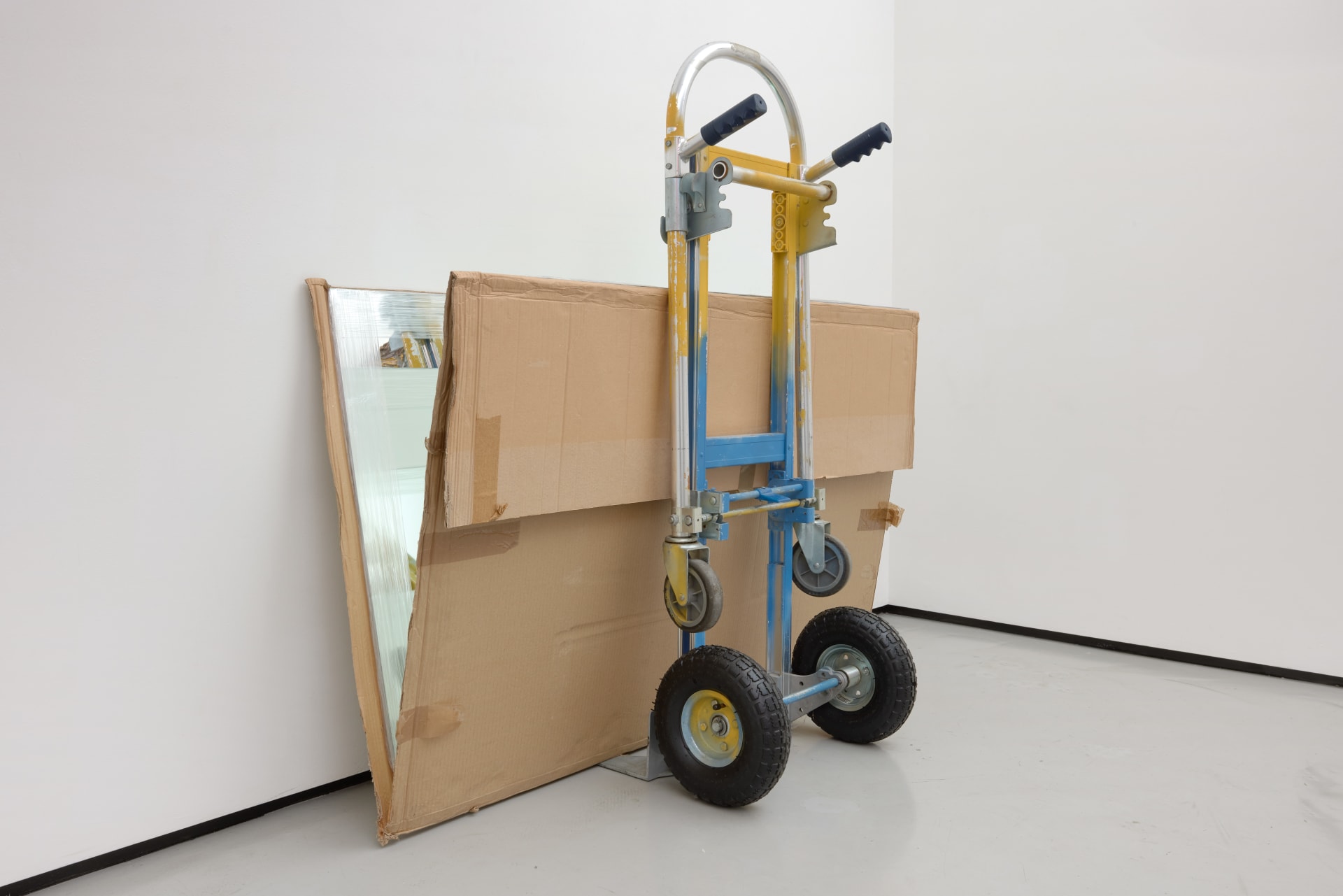 A sculpture by Rafael Pérez Evans consisting of a yellow and blue parcel truck resting against the wall with mirrors packaged in cardboard propped open at 15 degrees with yams, making an wheel of yams