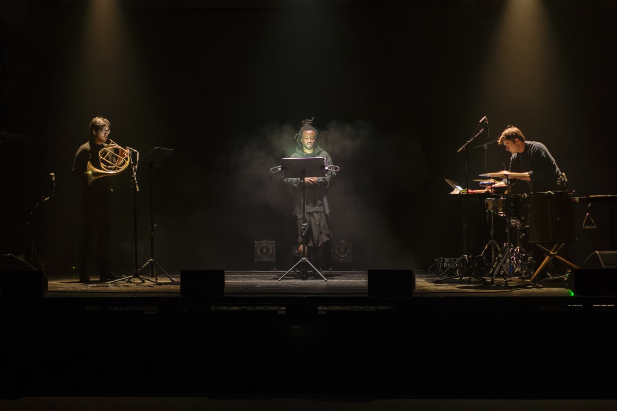 Simeon Barclay performing on a spotlit stage flanked by two musicians