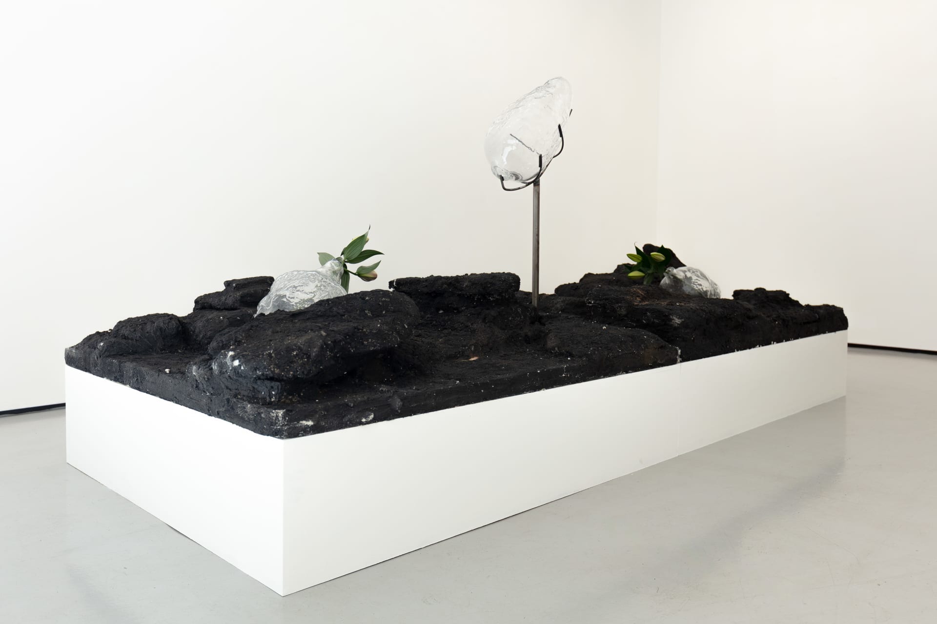 Blown glass vessels with lily flowers are held by metal structures above volcanic rock-like strata in this sculpture by Emii Alrai