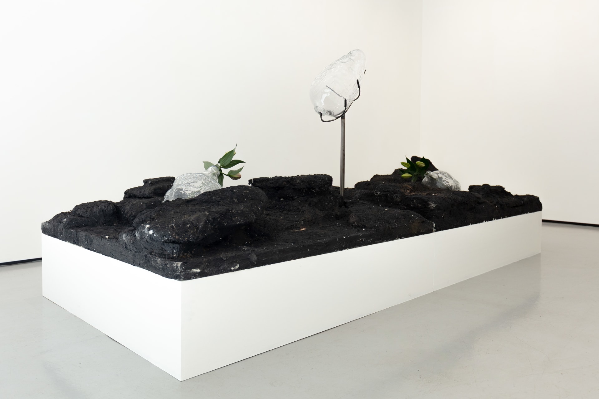 Blown glass vessels with lily flowers are held by metal structures above volcanic rock-like strata in this sculpture by Emii Alrai