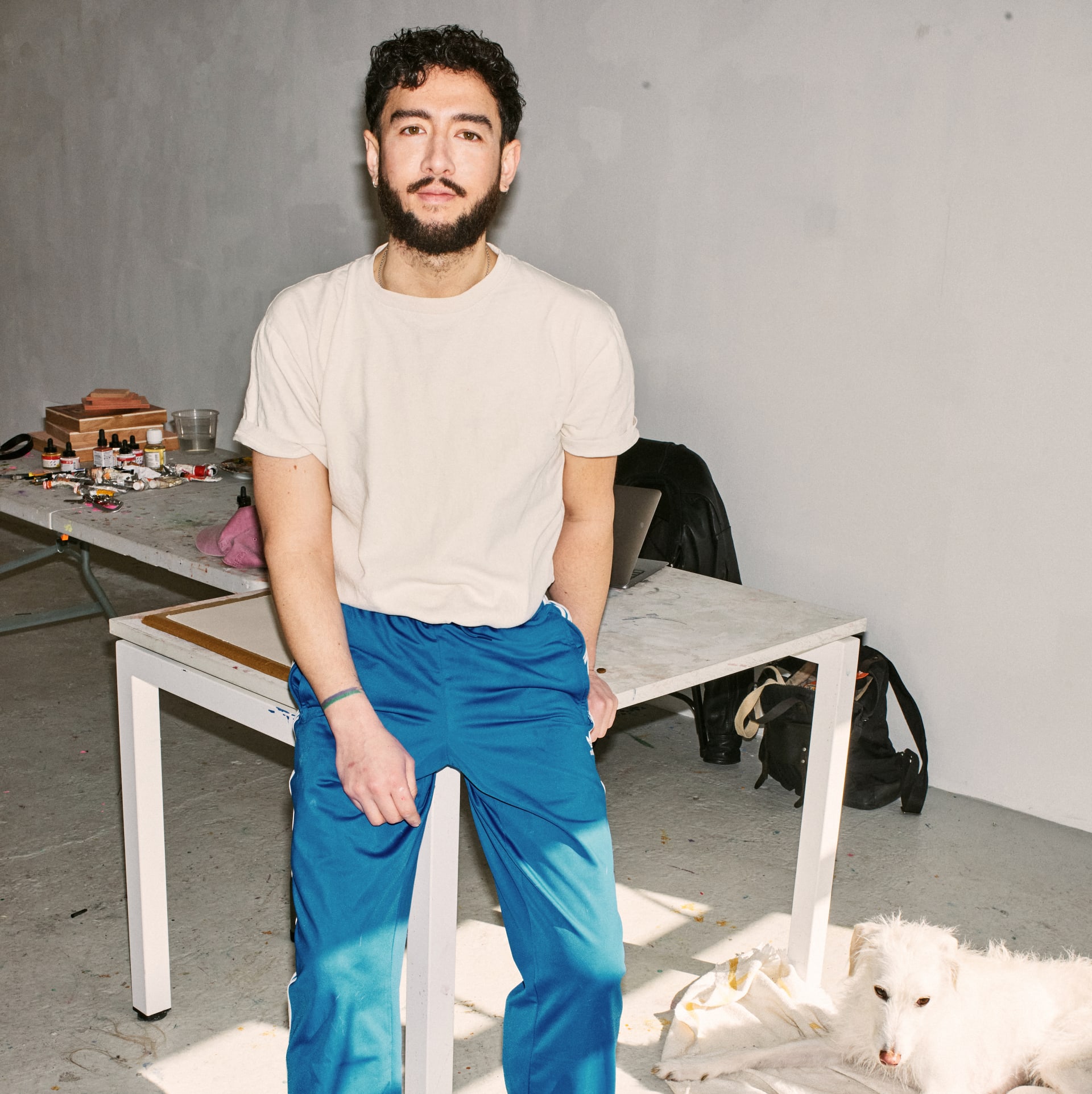 A portait of Ki Yoong leant on a table in his studio wearing a white t-shirt and ablue trousers