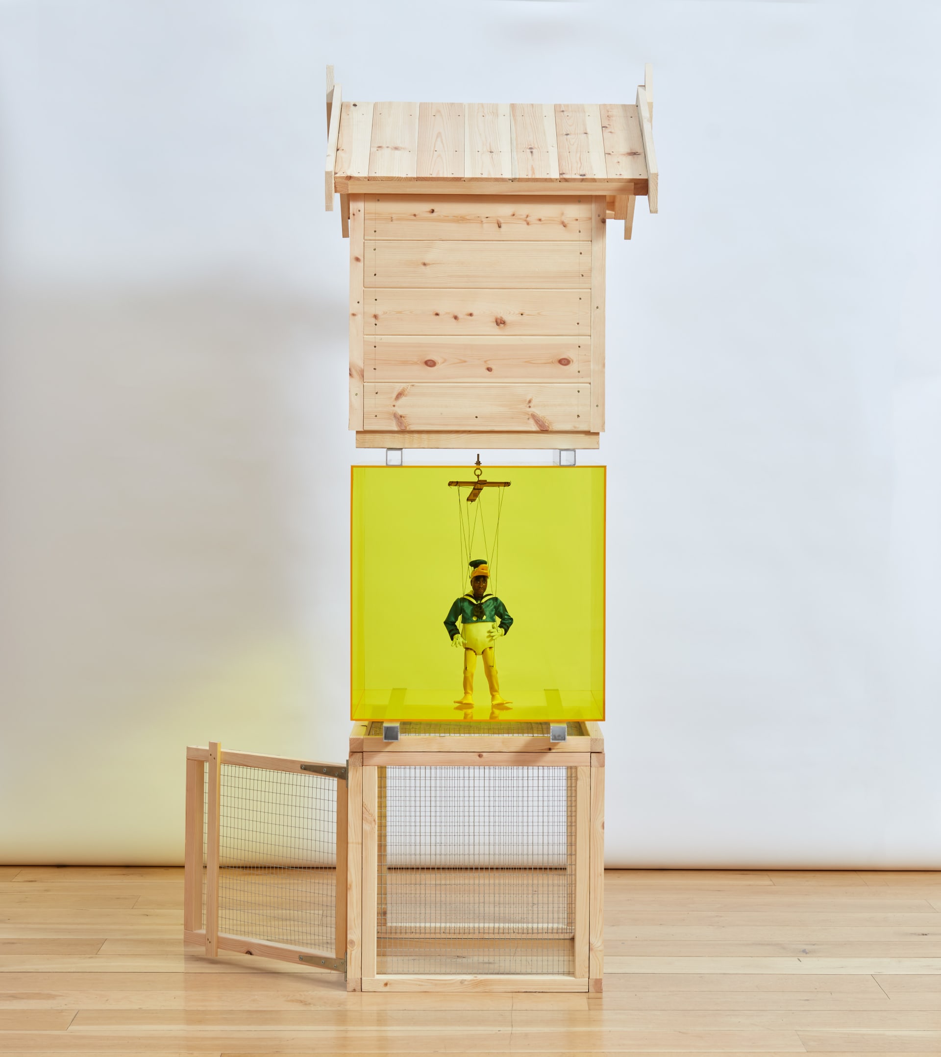 Iceberg is a sculpture by Simeon Barclay comprised of a stack of a hutch, a yellow perspex cube with a wooden puppet of the artist in Elton John's 'Donald Duck' costume and a dog kennel