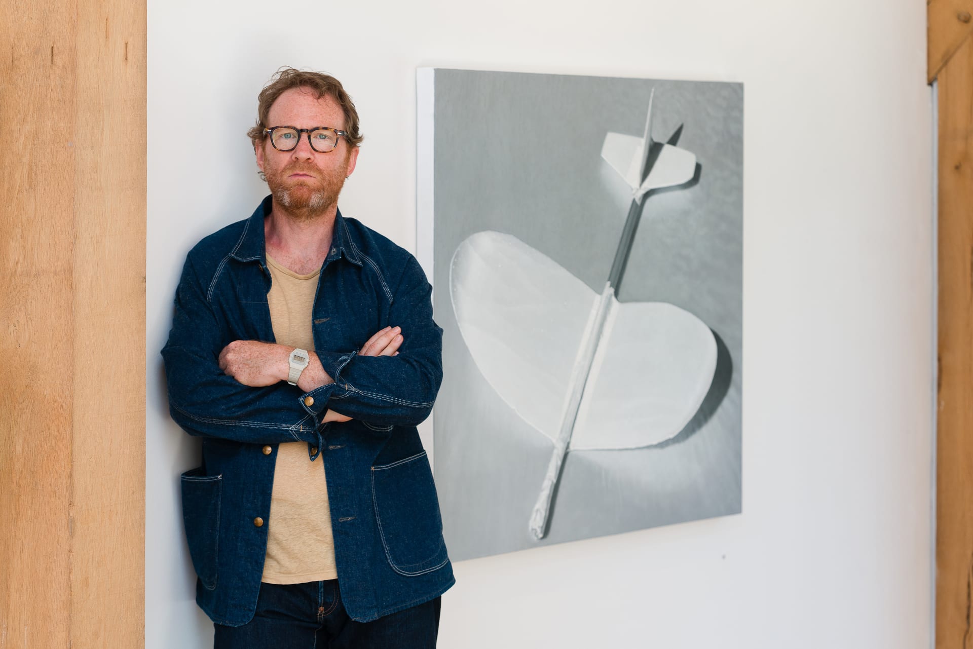 A portrait of Andrew Bryant stood in his studio in front of a monochrome painting of a toy plane