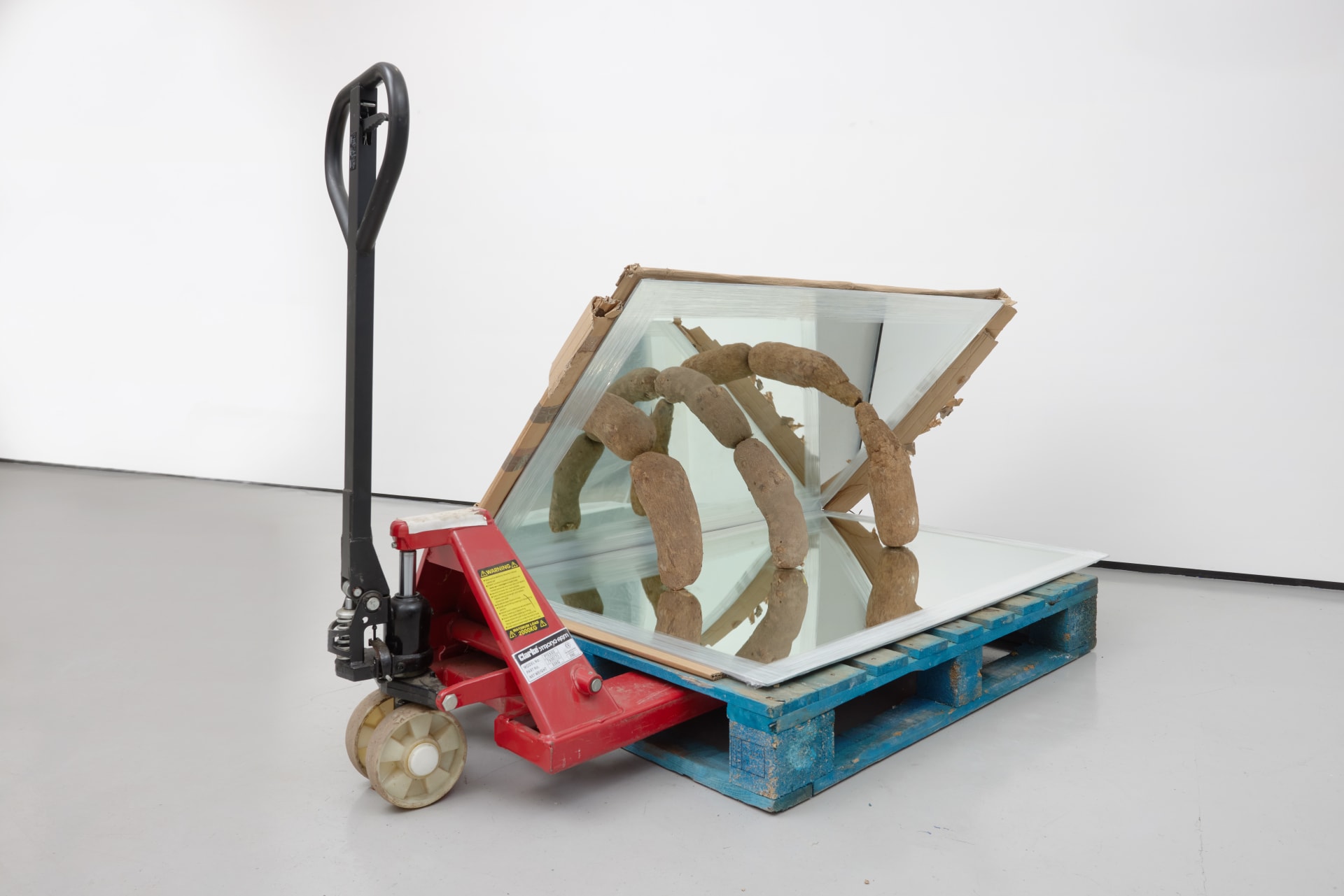 an image of a sculpture by Rafael Pérez Evans consisting of a pallet truck and pallet with two mirrors propped at a 45 degree angle by three yams, creating a wheel of yams effect 