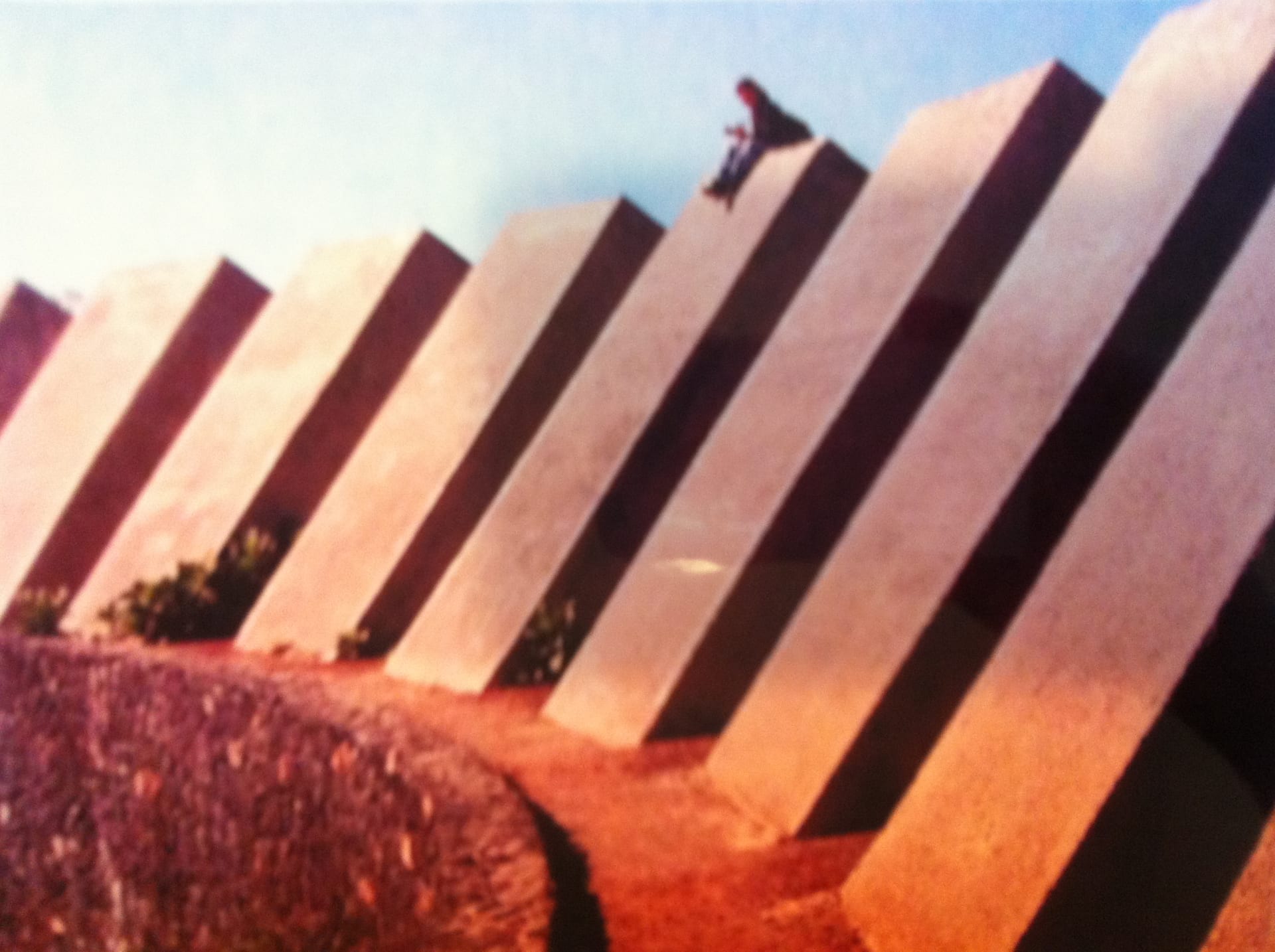 a blurred photograph by Cath Campbell of a video of a figure sat upon diagonal brutal architecture 