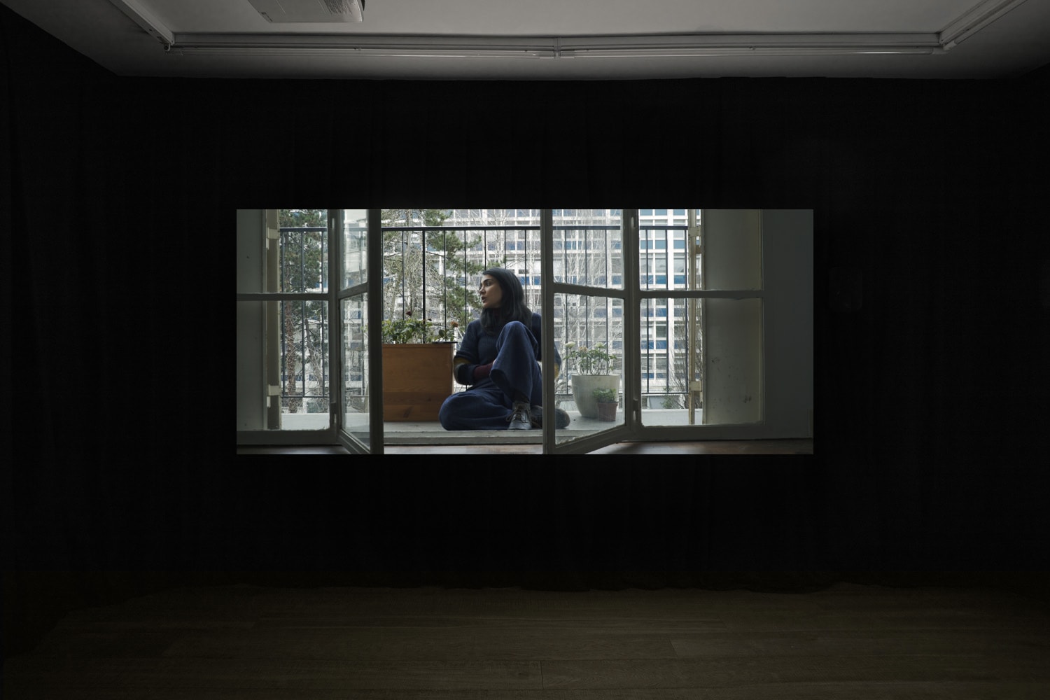 An installation image of Nia Fekri's film in Workplace's gallery in Margaret Street - a woman sits on a balcony of an urban apartment amidst potted plants. In the background is an apartment block