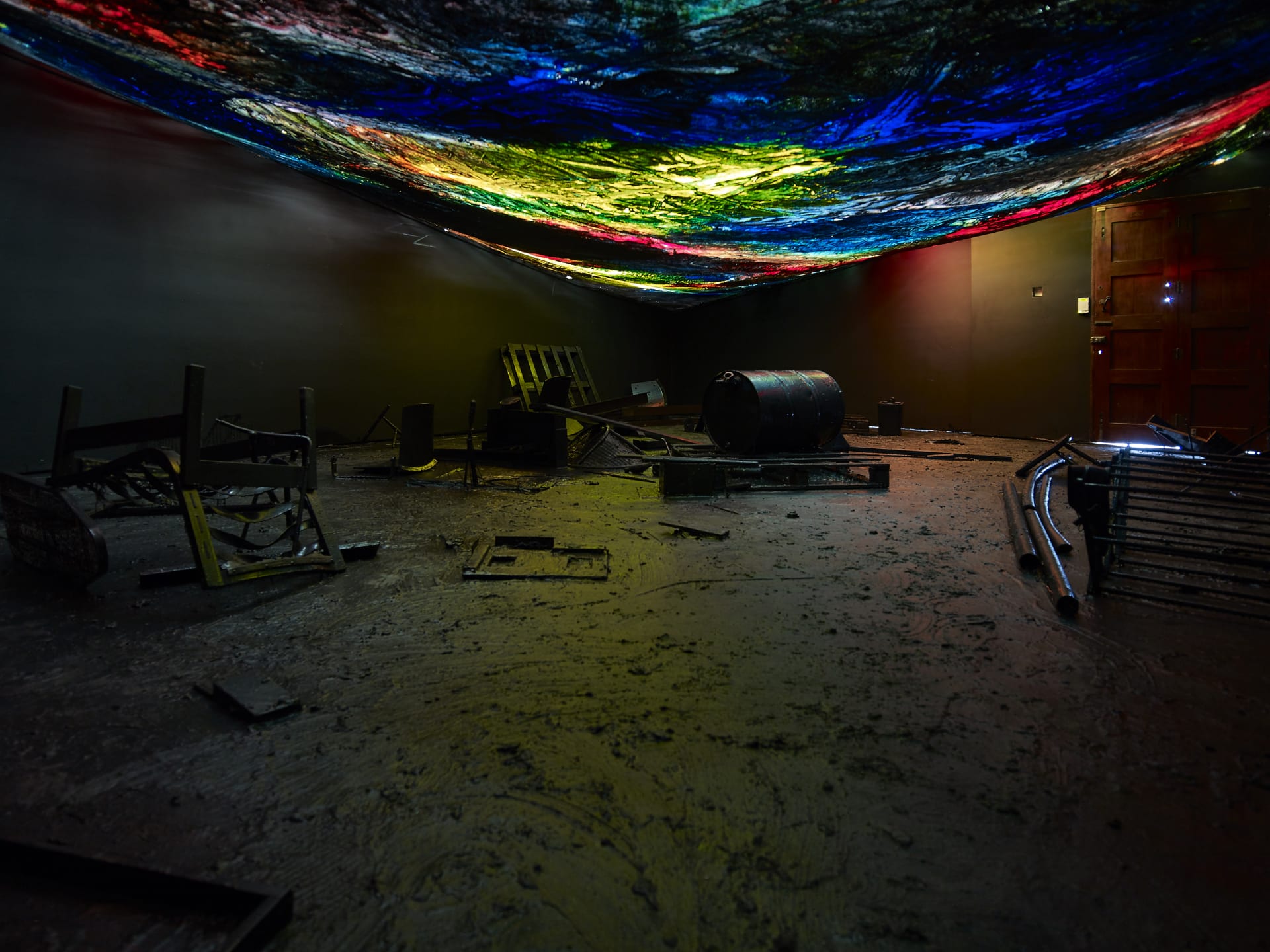 A installation by Hugo Canoilas consisting of darkened room with objects covered in black latex, and a low painted colourful canvas as ceiling