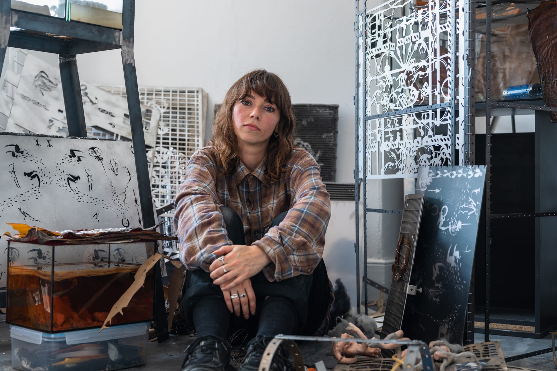 A woman with brown hair sat on the floor looking directly to camera surrounded by numerous laser-cut metal panels
