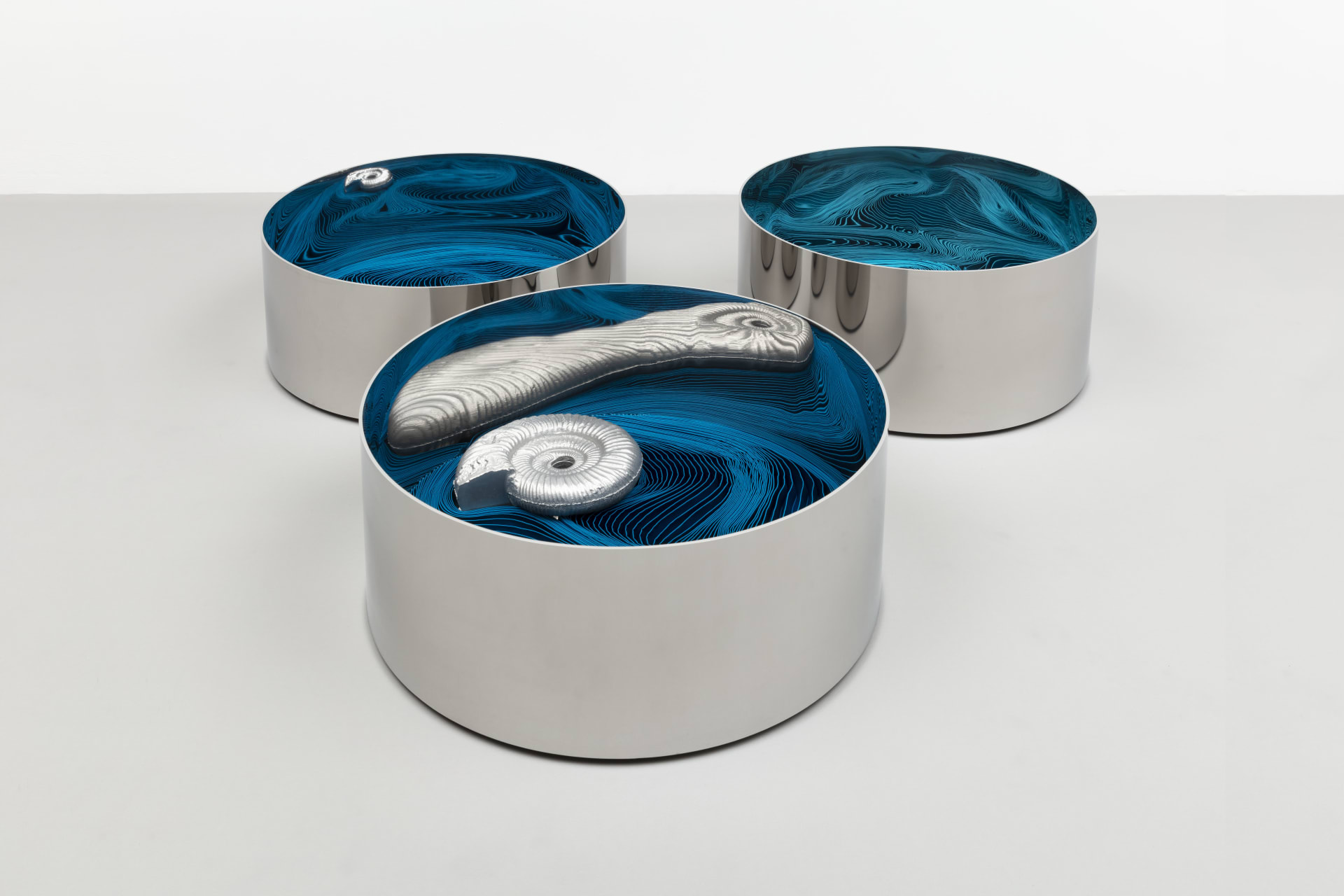 Sculpture by Alice Channer, three steel casks with pleated turquoise fabric inside and casts of an ammonite sitting on top