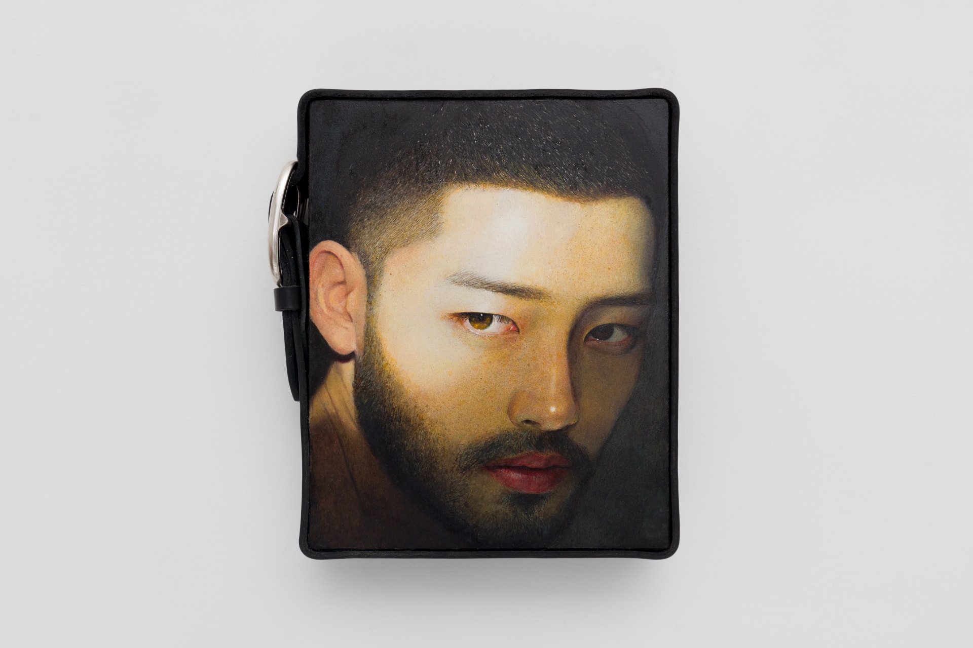A portrait by Ki Yoong of a young Asian man looking at us with a shaft of light across his face. A belt with a large buckle is around the painting