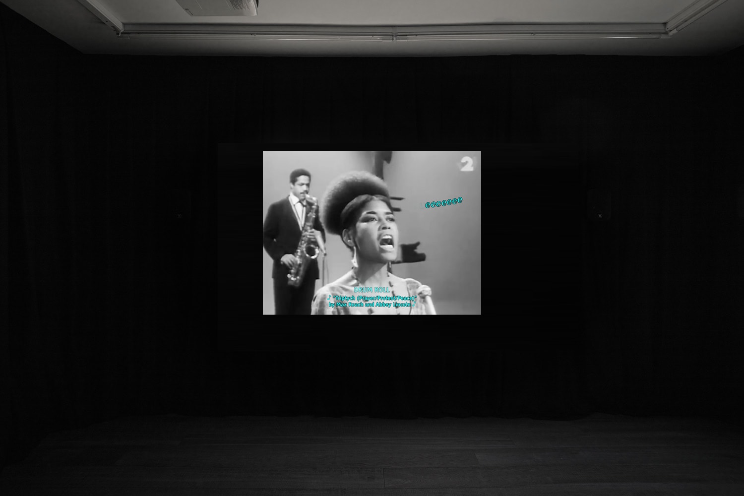 An install shot of Rosa-Johan Uddoh's film at Workplace in London. A black and white image of a saxophonist and singer performing. Over the top is blue text 'eeeeeee' describing the sound.