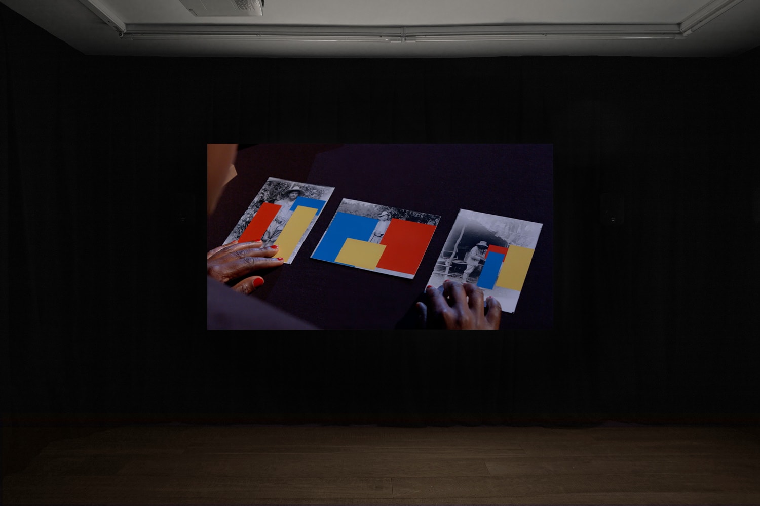 An installation image of Belinda Kazeem-Kaminski's film in Workplace's gallery in Margaret Street - the camera looks over the shoulder of a black woman with red painted fingernails looking at printed images of black and white images of people covered over with brightly coloured rectangles