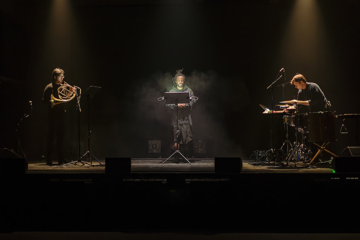 Simeon Barclay performing with two musicians on a dark stage